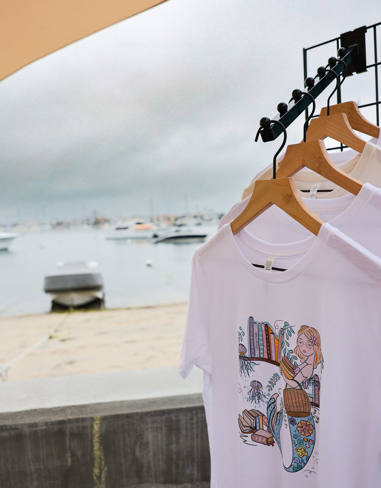 Illustrated graphic of a mermaid librarian on a white cotton t-shirt, surrounded by books and ocean creatures, with a friendly smile and a tail instead of legs, designed for youth sizes