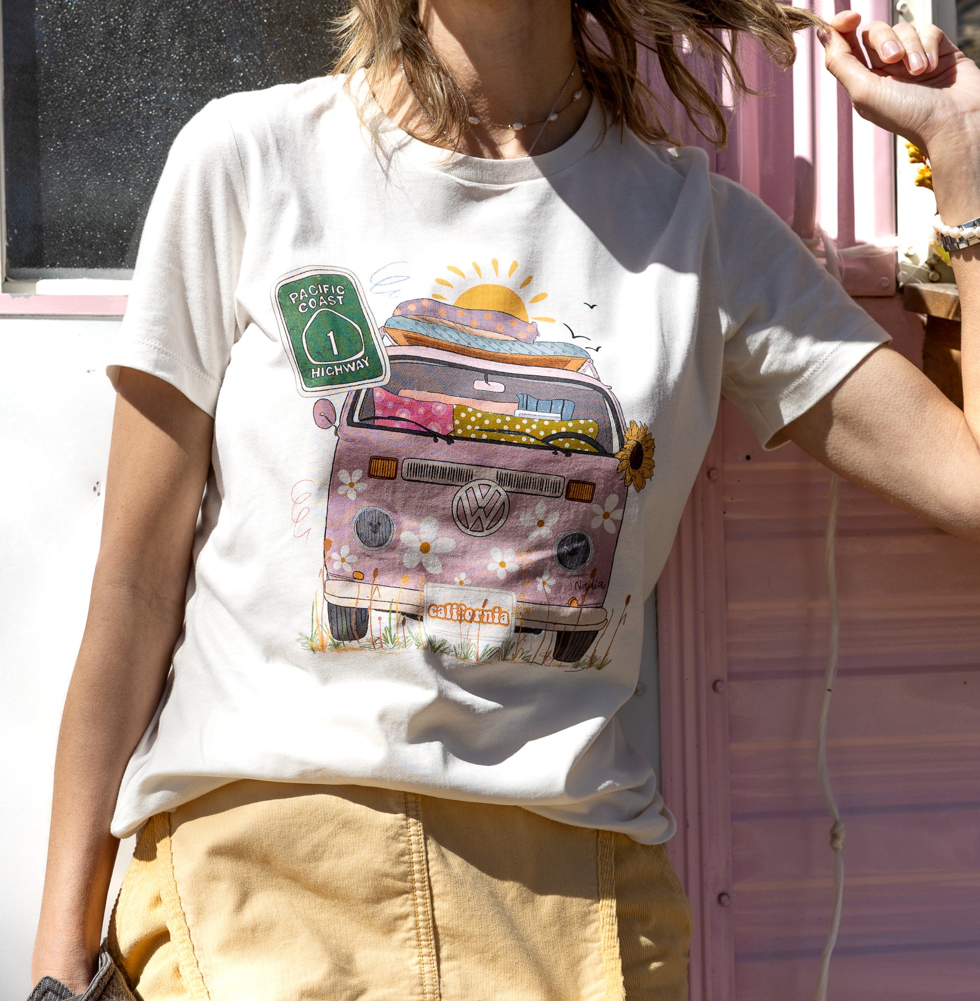 Hand-illustrated graphic tee featuring a vibrant pink and floral Hippie bus with California license plate and Highway 1 sign.
