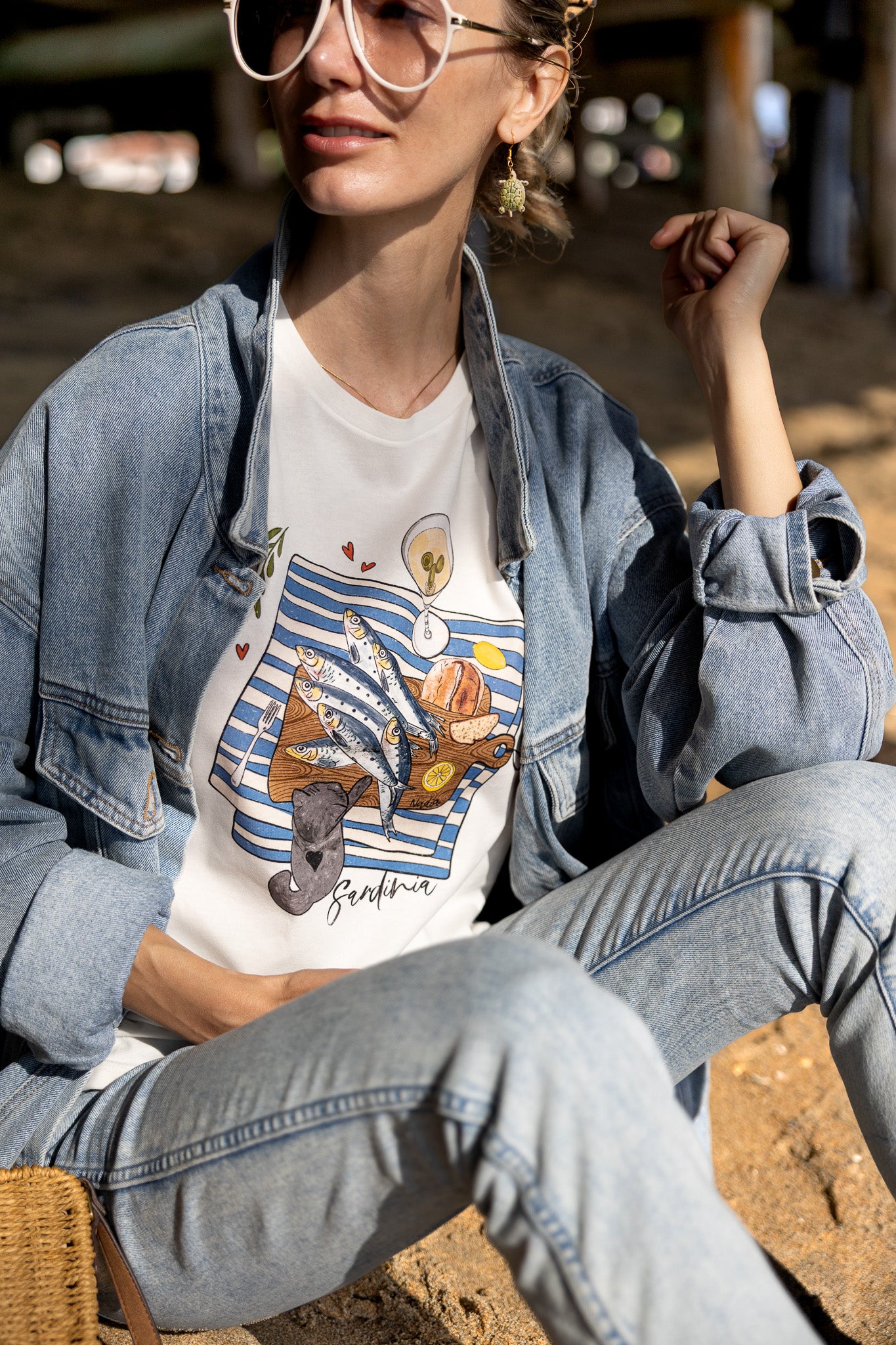 Woman is wearing a graphic tee with hand-illustrated sardines on a cutting board, dirty martini, bread, lemon, and a cute cat reaching for sardines. SARDINIA slogan