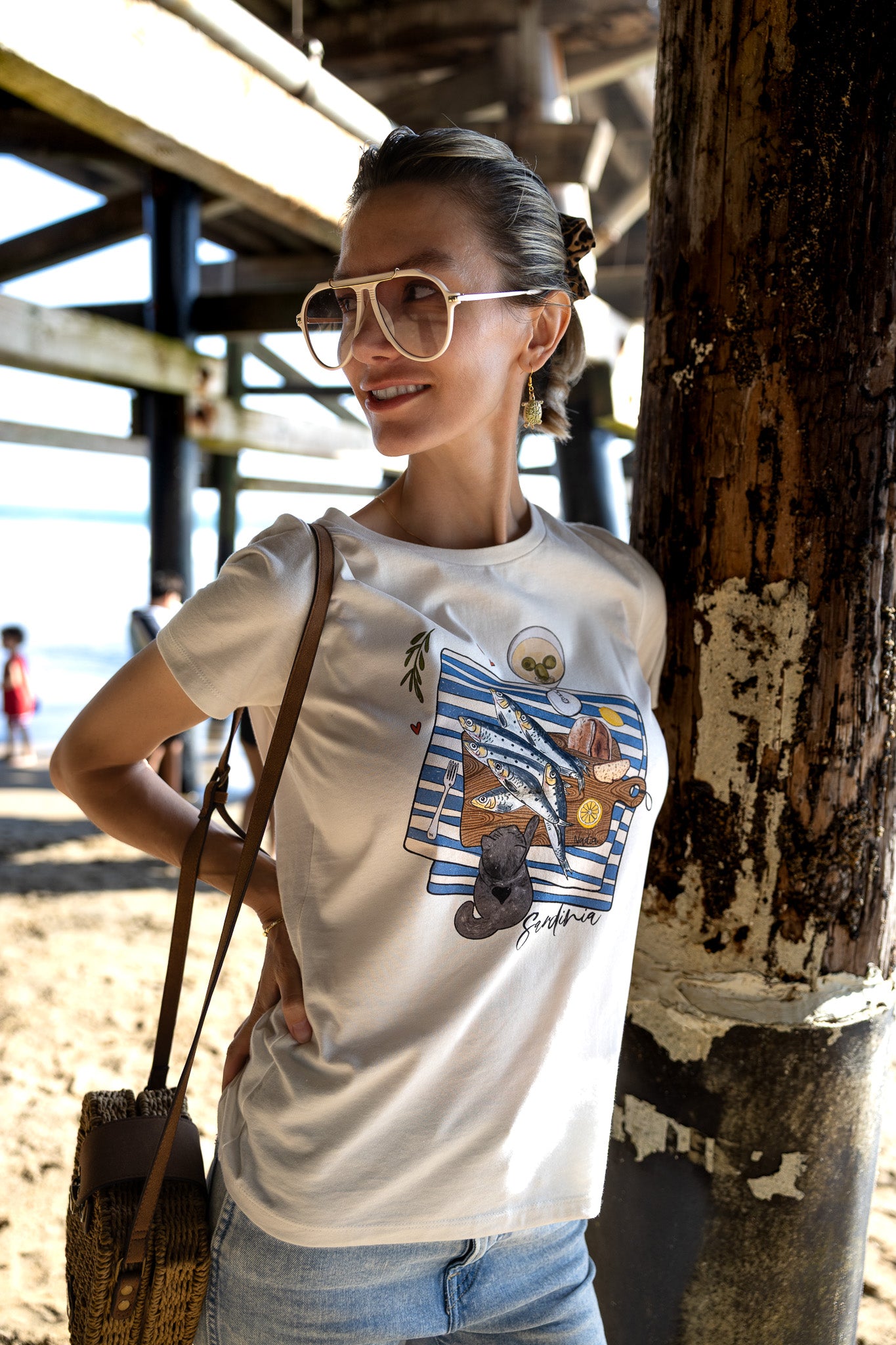 Woman is wearing a graphic tee with hand-illustrated sardines on a cutting board, dirty martini, bread, lemon, and a cute cat reaching for sardines. SARDINIA slogan