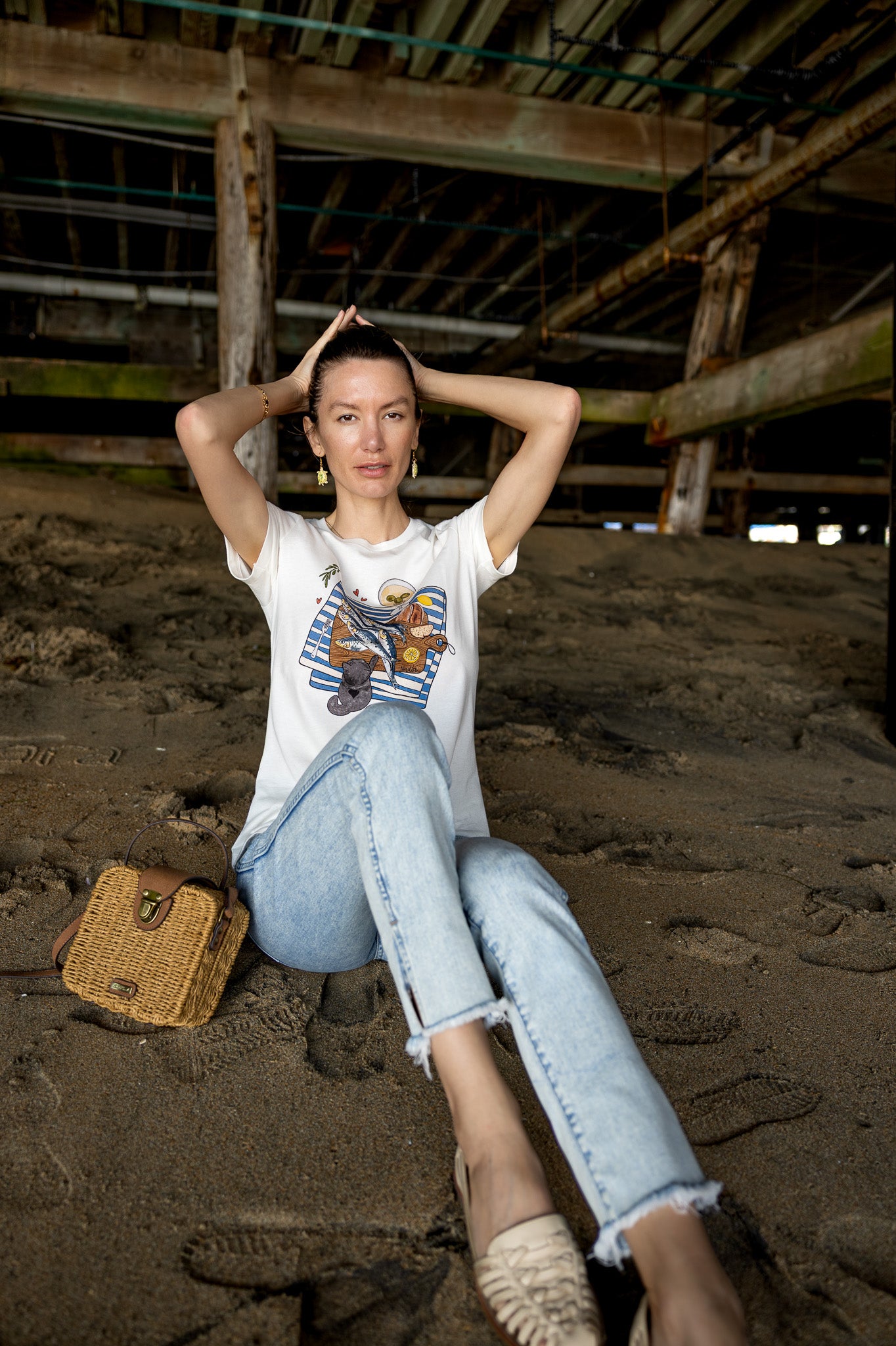 Woman is wearing a graphic tee with hand-illustrated sardines on a cutting board, dirty martini, bread, lemon, and a cute cat reaching for sardines. SARDINIA slogan