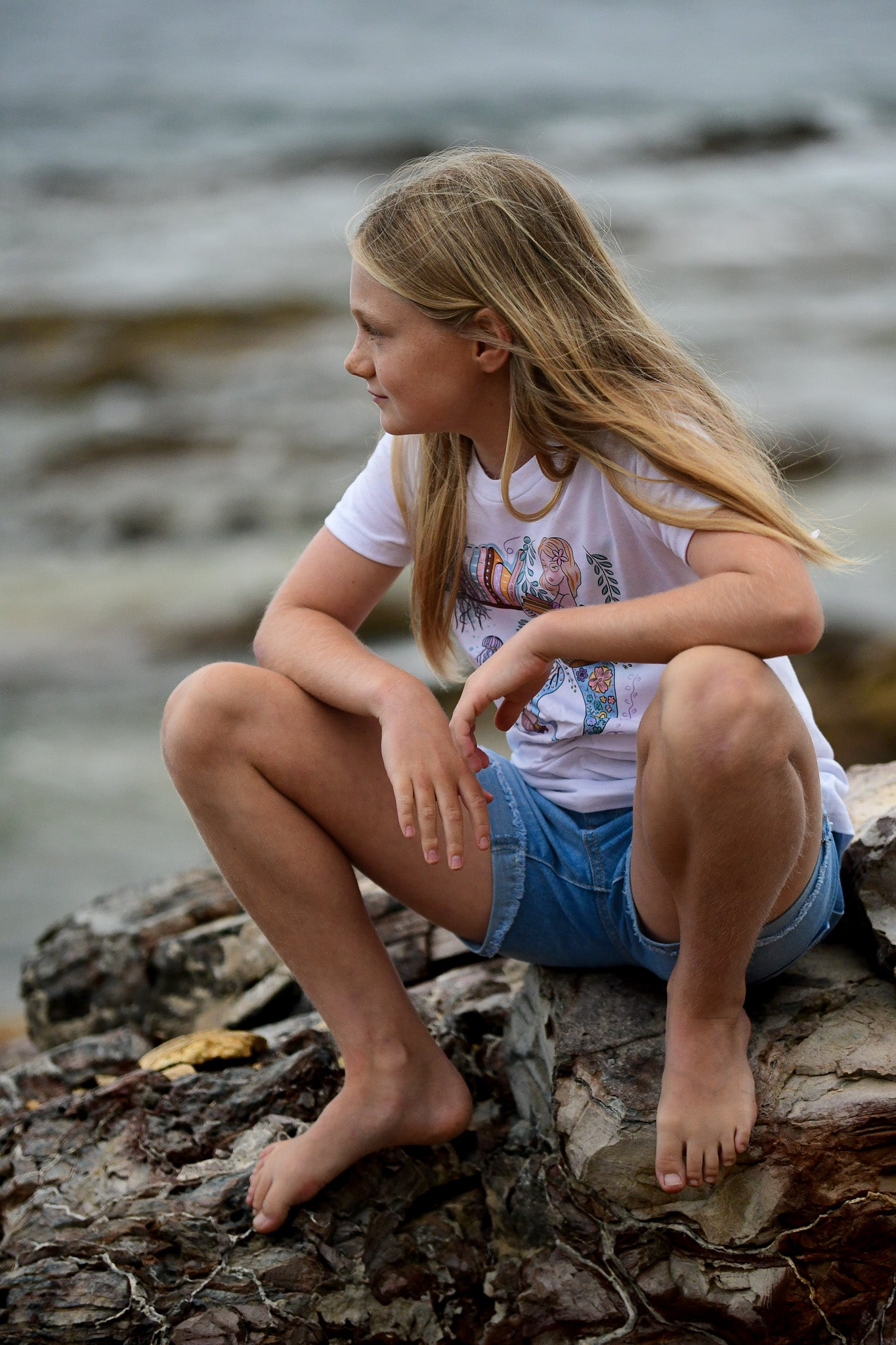 Illustrated graphic of a mermaid librarian on a white cotton t-shirt, surrounded by books and ocean creatures, with a friendly smile and a tail instead of legs, designed for youth sizes