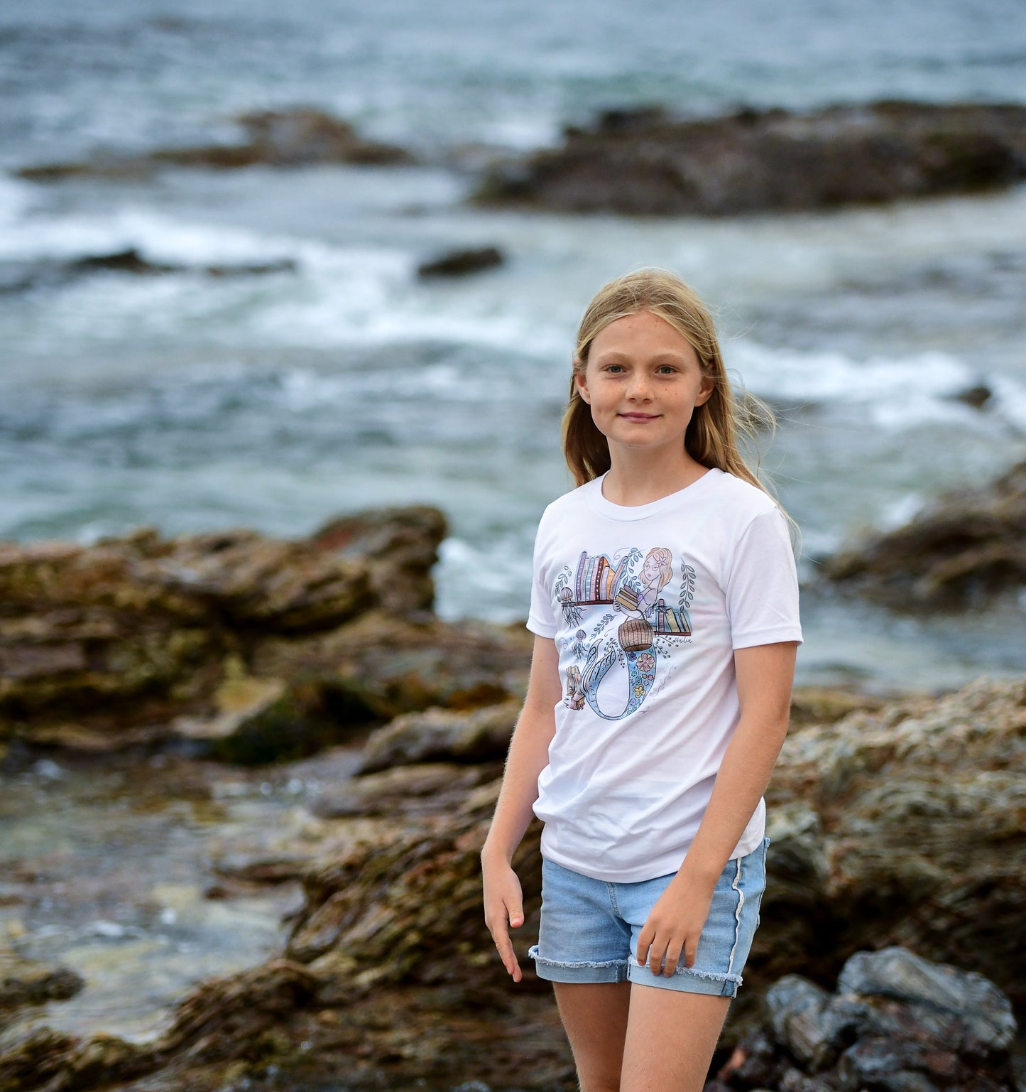 Illustrated graphic of a mermaid librarian on a white cotton t-shirt, surrounded by books and ocean creatures, with a friendly smile and a tail instead of legs, designed for youth sizes