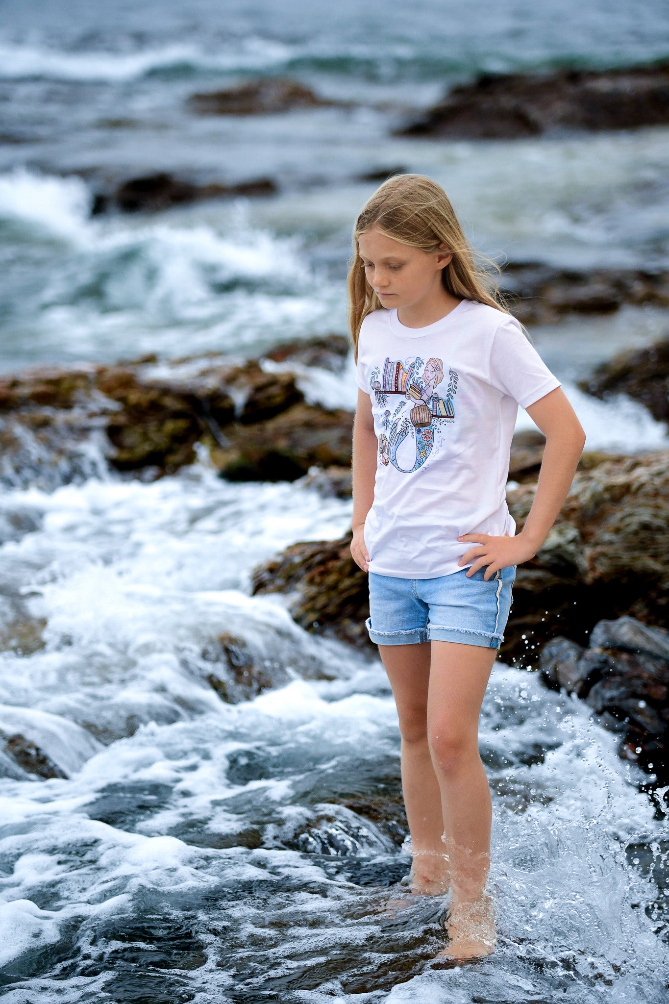 Illustrated graphic of a mermaid librarian on a white cotton t-shirt, surrounded by books and ocean creatures, with a friendly smile and a tail instead of legs, designed for youth sizes
