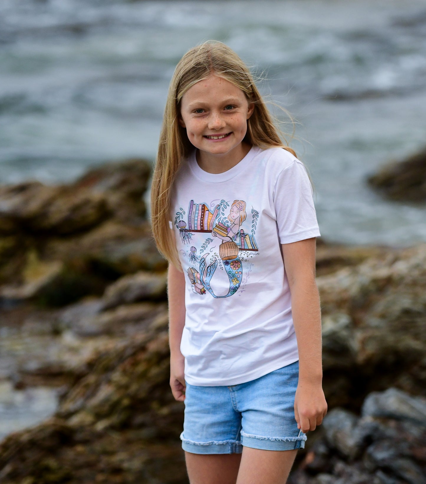 Illustrated graphic of a mermaid librarian on a white cotton t-shirt, surrounded by books and ocean creatures, with a friendly smile and a tail instead of legs, designed for youth sizes