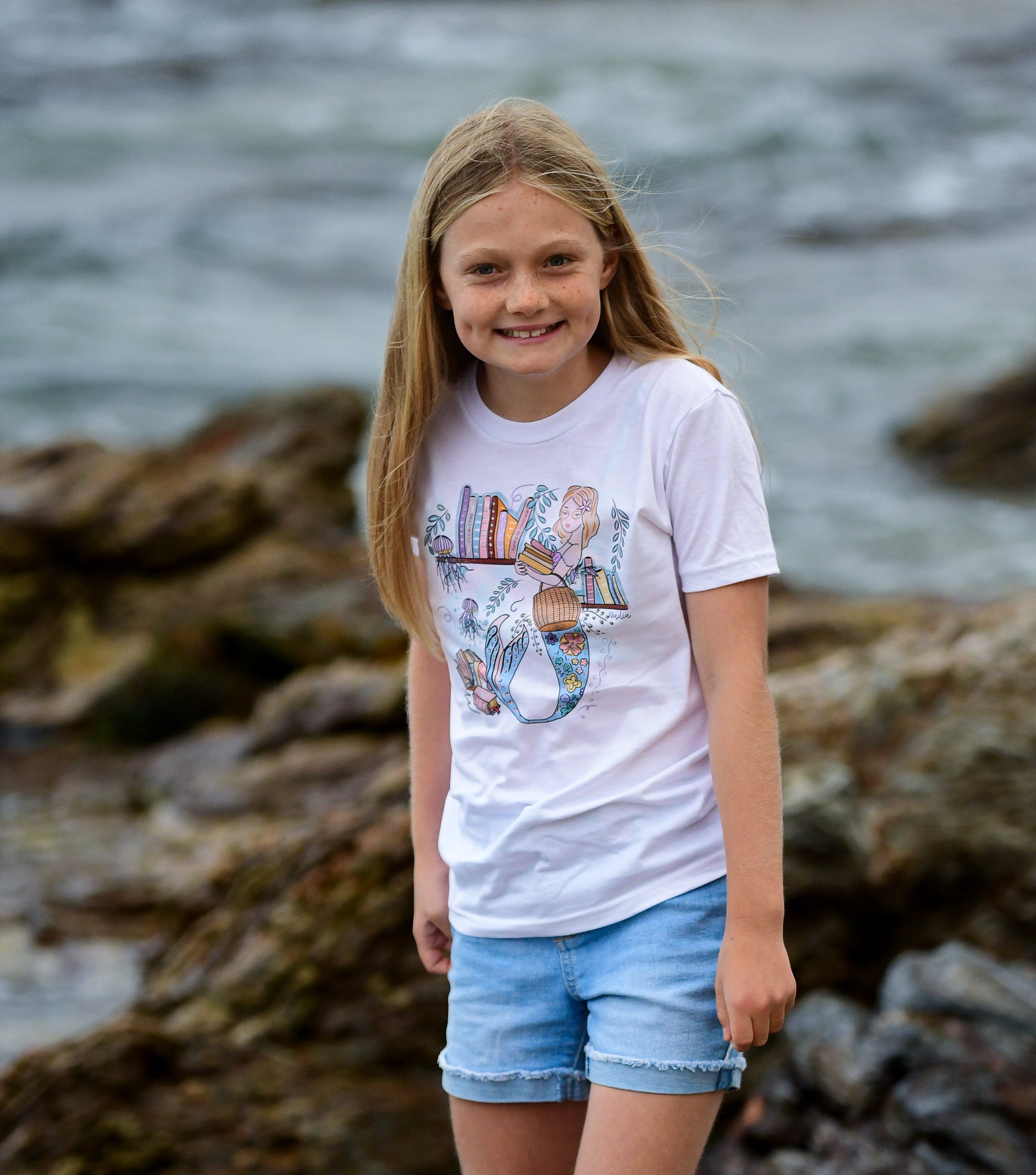 Illustrated graphic of a mermaid librarian on a white cotton t-shirt, surrounded by books and ocean creatures, with a friendly smile and a tail instead of legs, designed for youth sizes