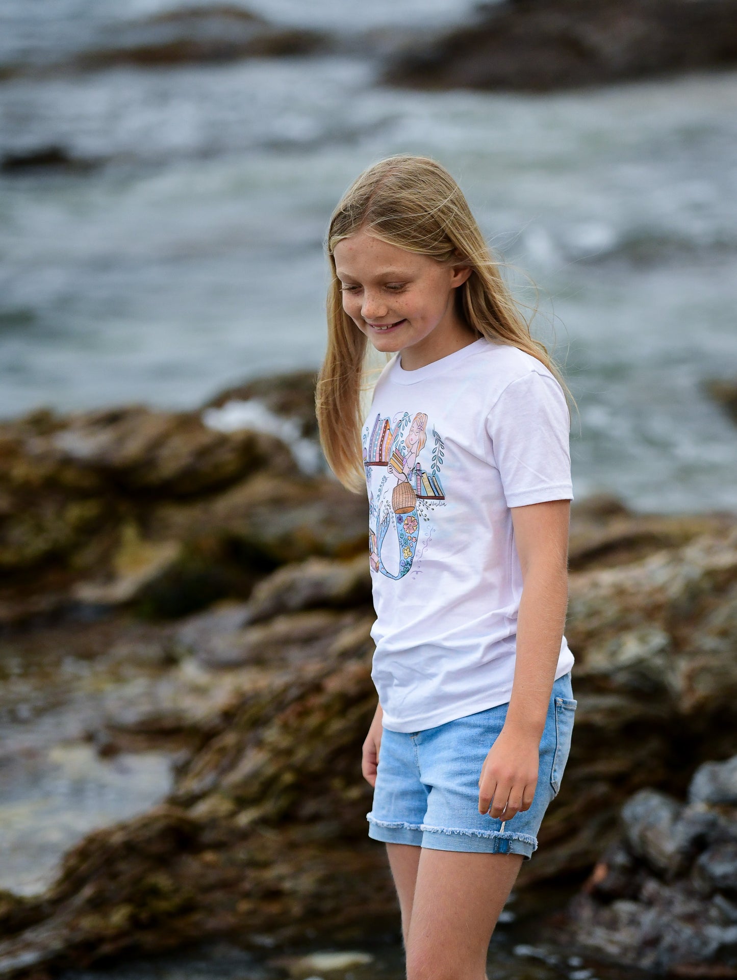 Illustrated graphic of a mermaid librarian on a white cotton t-shirt, surrounded by books and ocean creatures, with a friendly smile and a tail instead of legs, designed for youth sizes