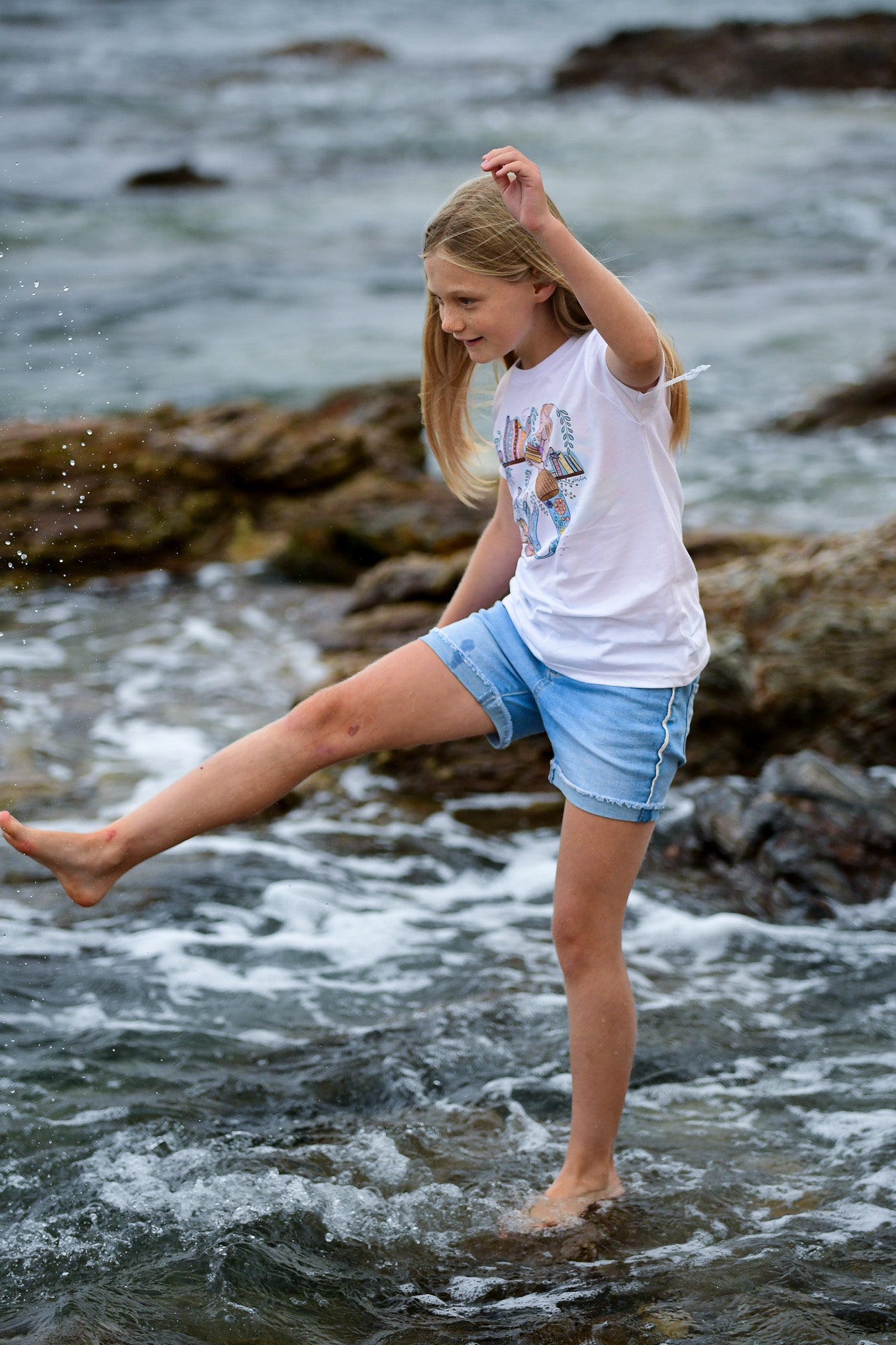 Illustrated graphic of a mermaid librarian on a white cotton t-shirt, surrounded by books and ocean creatures, with a friendly smile and a tail instead of legs, designed for youth sizes