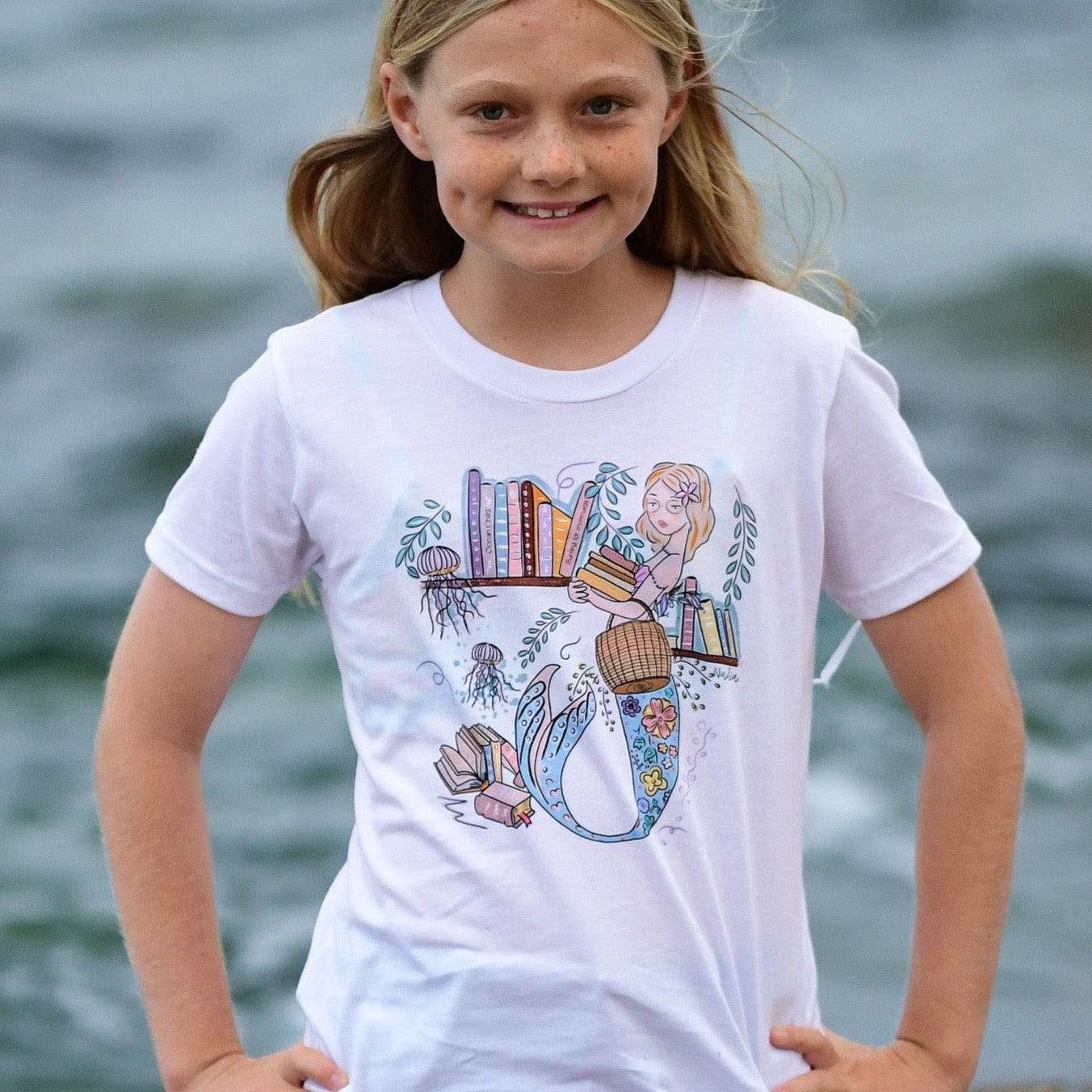 Illustrated graphic of a mermaid librarian on a white cotton t-shirt, surrounded by books and ocean creatures, with a friendly smile and a tail instead of legs, designed for youth sizes