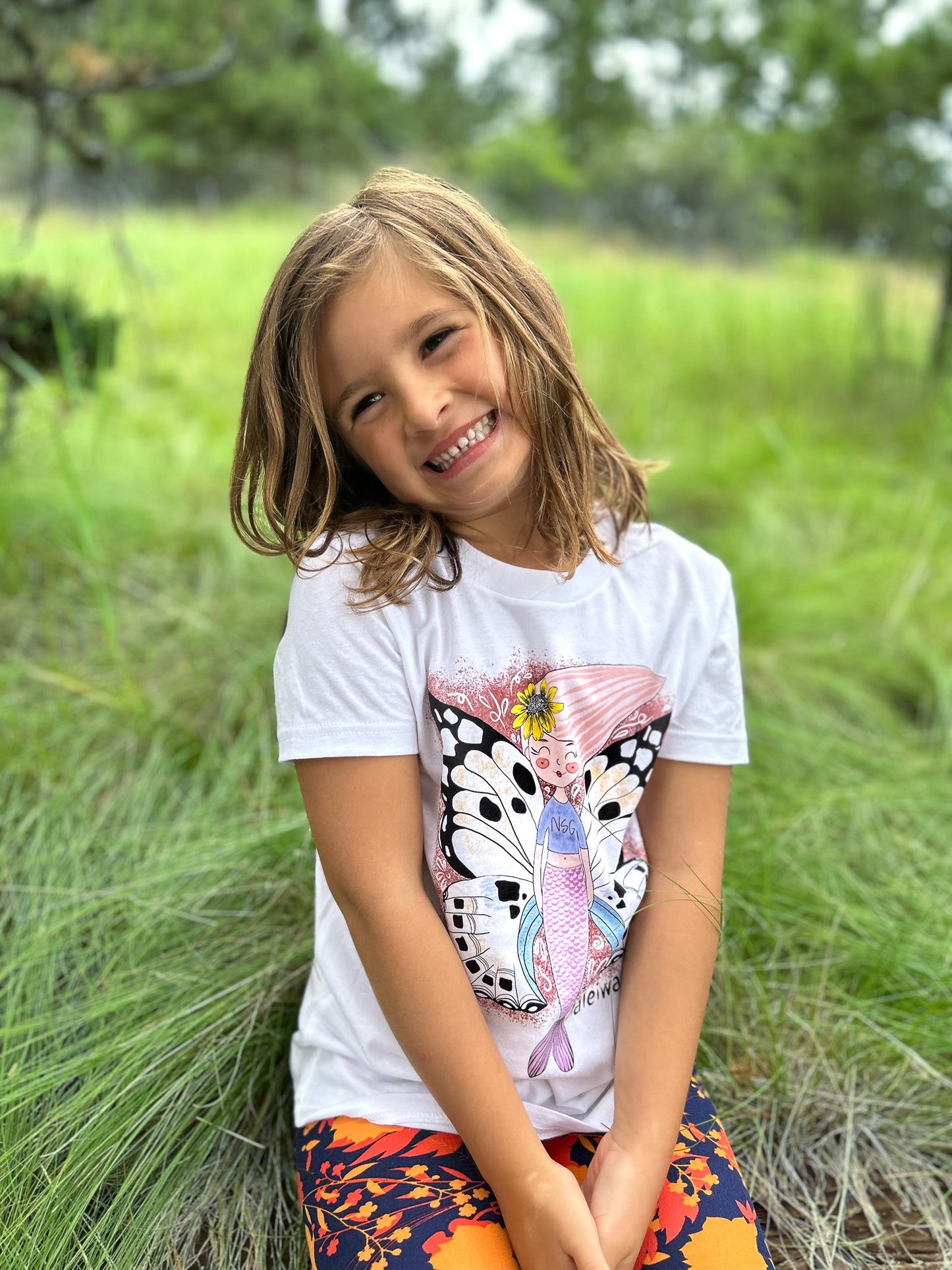 Illustrated graphic of a mermaid with colorful butterfly wings, surrounded by tropical flowers and leaves. The mermaid has a smiling face and long, flowing hair. The design is printed on a white toddler t-shirt.