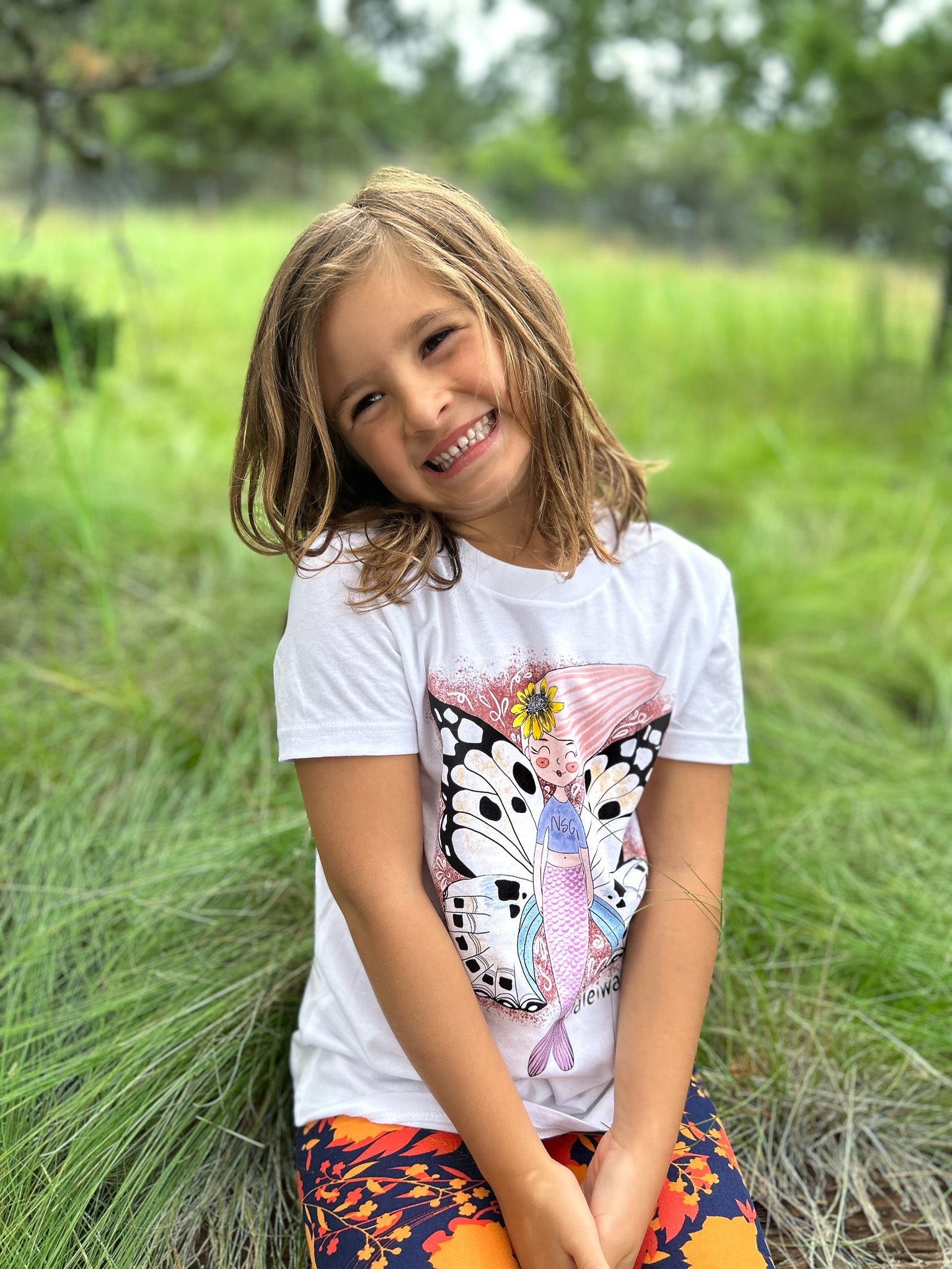 Illustrated graphic of a mermaid with colorful butterfly wings, surrounded by tropical flowers and leaves. The mermaid has a smiling face and long, flowing hair. The design is printed on a white toddler t-shirt.