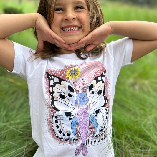 Illustrated graphic of a mermaid with colorful butterfly wings, surrounded by tropical flowers and leaves. The mermaid has a smiling face and long, flowing hair. The design is printed on a white toddler t-shirt.