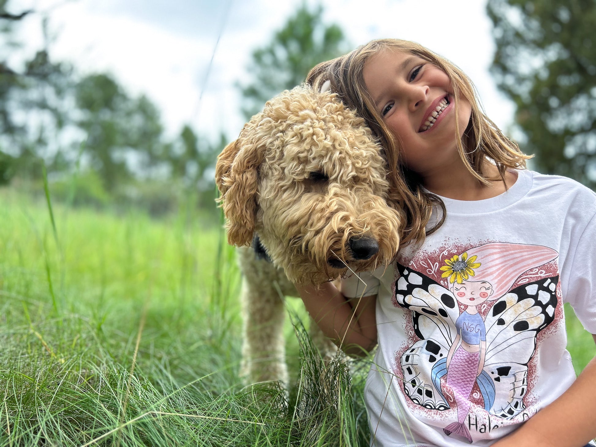 Illustrated graphic of a mermaid with colorful butterfly wings, surrounded by tropical flowers and leaves. The mermaid has a smiling face and long, flowing hair. The design is printed on a white toddler t-shirt.