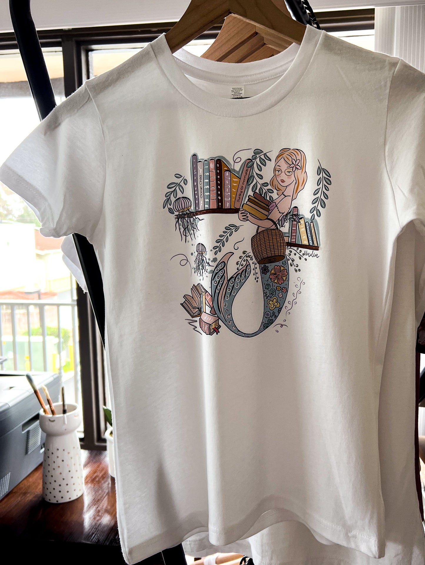 Illustrated graphic of a mermaid librarian on a white cotton t-shirt, surrounded by books and ocean creatures, with a friendly smile and a tail instead of legs, designed for youth sizes