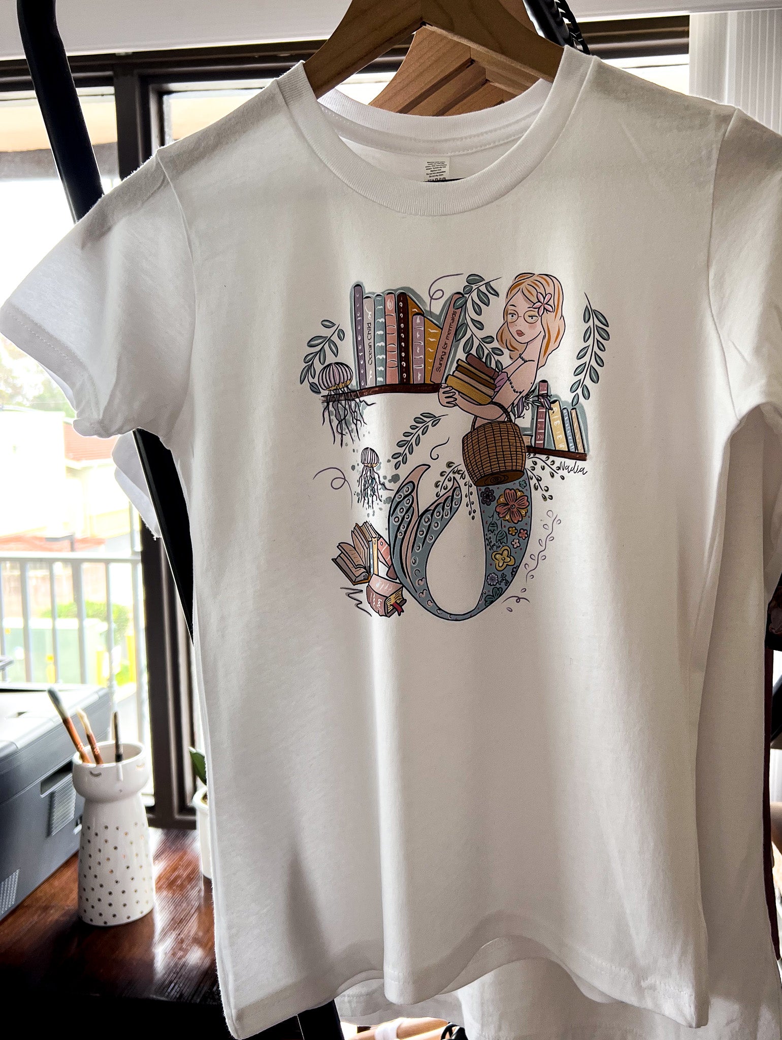 Illustrated graphic of a mermaid librarian on a white cotton t-shirt, surrounded by books and ocean creatures, with a friendly smile and a tail instead of legs, designed for youth sizes