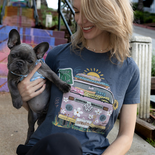 Woman in a blue North Shore Girls graphic t-shirt holding a puppy on colorful stairs; the shirt features a hand-drawn vintage floral beach van.