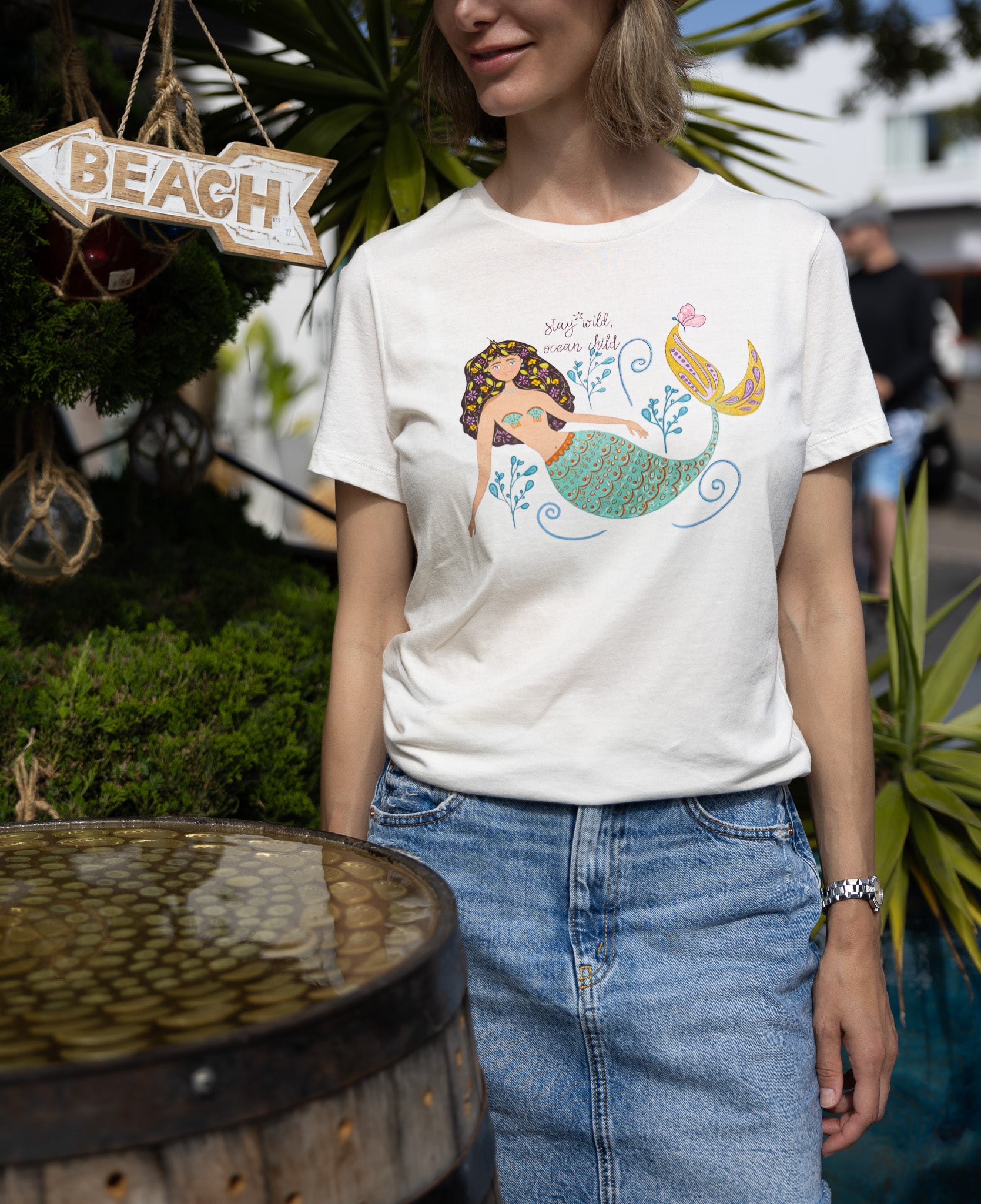 Illustrated mermaid graphic tee for women with 'Stay Wild Ocean Child' slogan. A whimsical mermaid tail and sea flowers surround the text, creating a dreamy ocean-inspired design on a relaxed-fit cotton t-shirt.