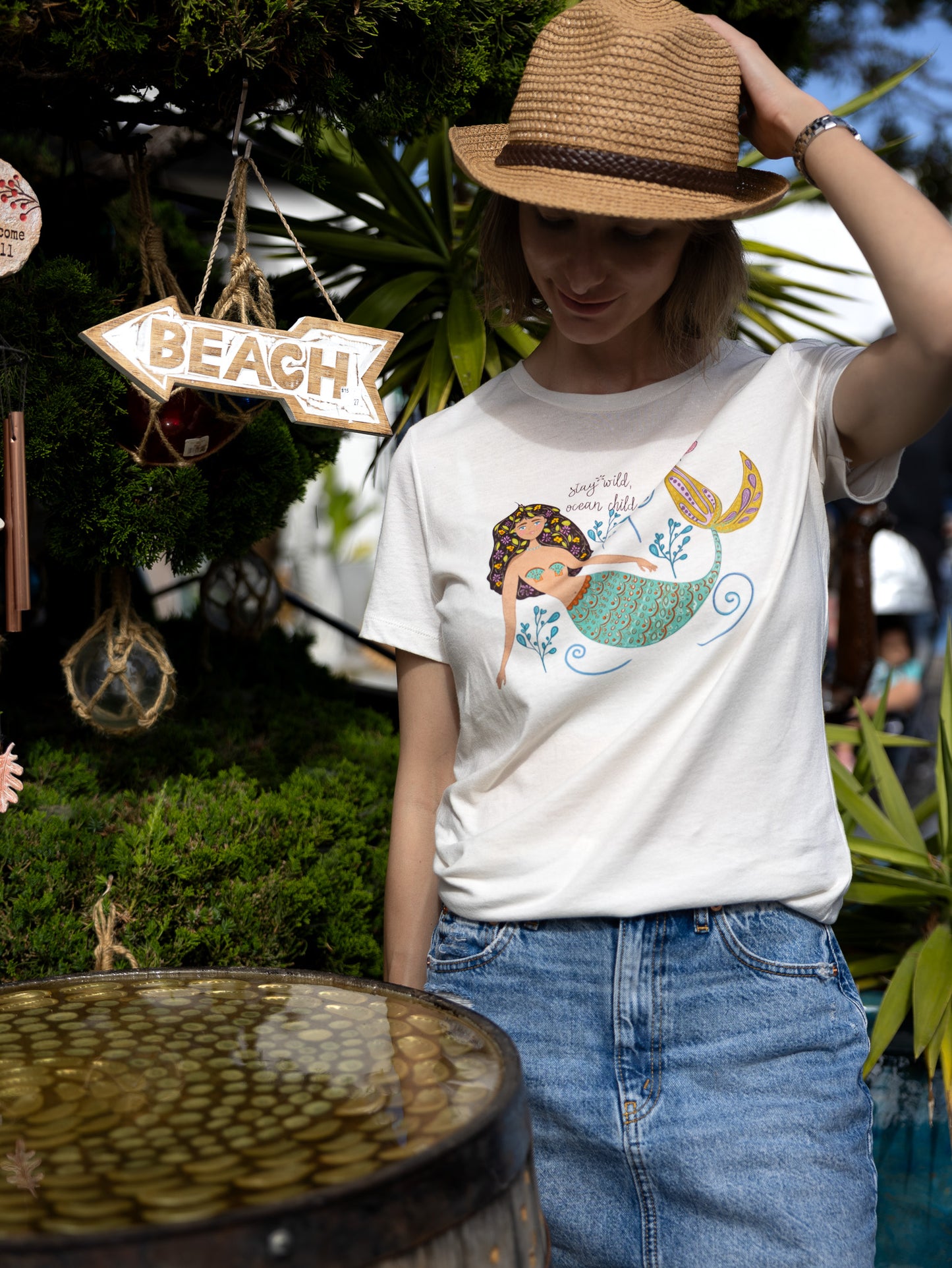 Illustrated mermaid graphic tee for women with 'Stay Wild Ocean Child' slogan. A whimsical mermaid tail and sea flowers surround the text, creating a dreamy ocean-inspired design on a relaxed-fit cotton t-shirt.