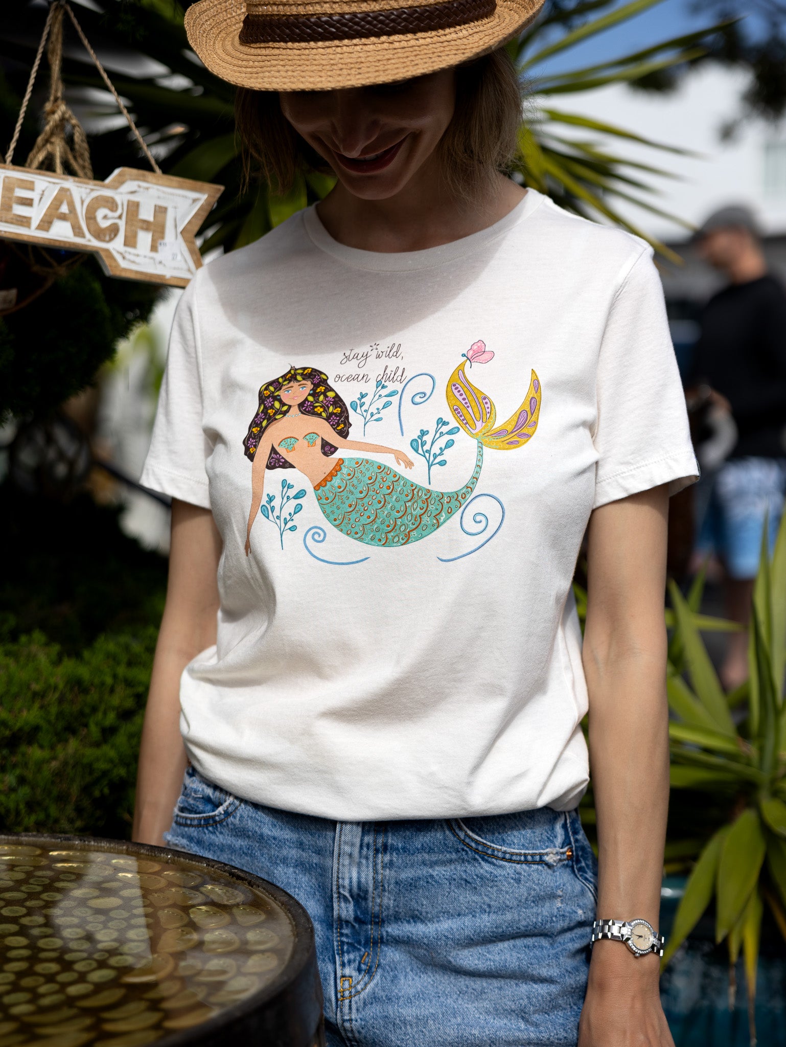 Illustrated mermaid graphic tee for women with 'Stay Wild Ocean Child' slogan. A whimsical mermaid tail and sea flowers surround the text, creating a dreamy ocean-inspired design on a relaxed-fit cotton t-shirt.