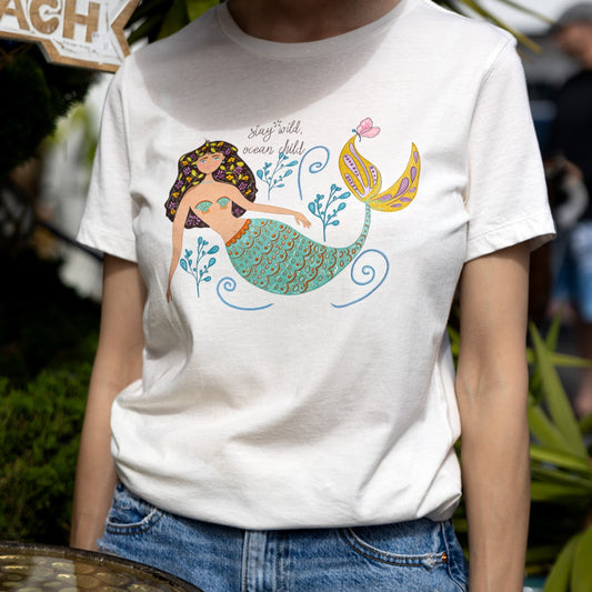 Illustrated mermaid graphic tee for women with 'Stay Wild Ocean Child' slogan. A whimsical mermaid tail and sea flowers surround the text, creating a dreamy ocean-inspired design on a relaxed-fit cotton t-shirt.