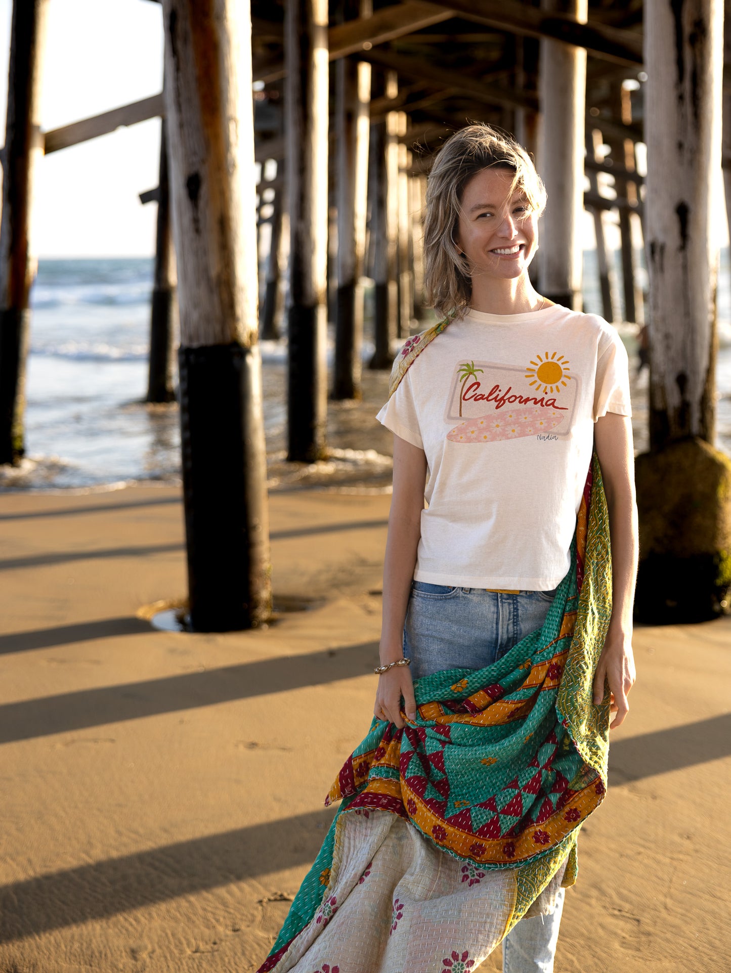 Relaxed fit women's graphic tee with hand-drawn California design, featuring a pink floral surfboard, palm tree, and sun on a white background