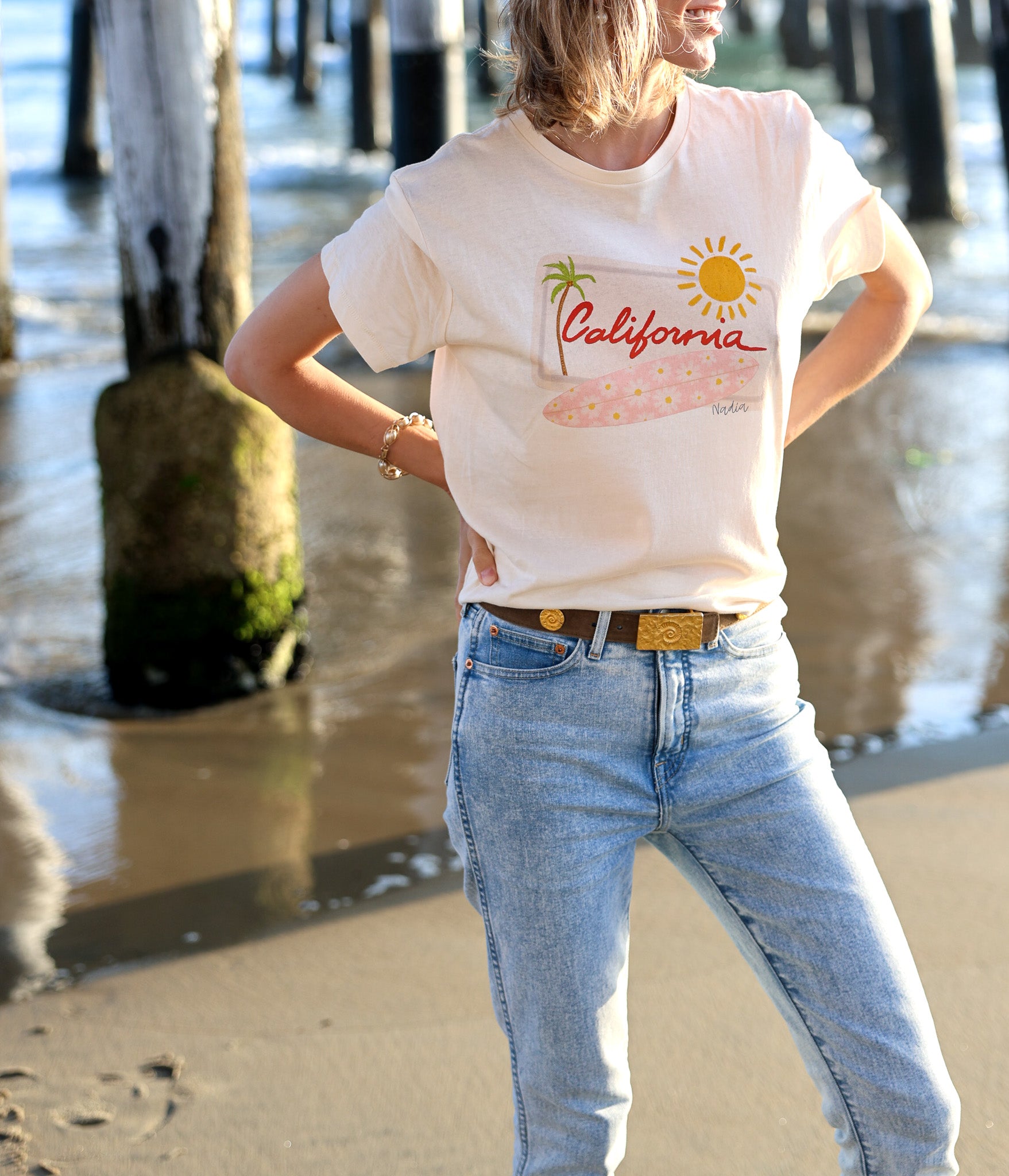 Relaxed fit women's graphic tee with hand-drawn California design, featuring a pink floral surfboard, palm tree, and sun on a white background