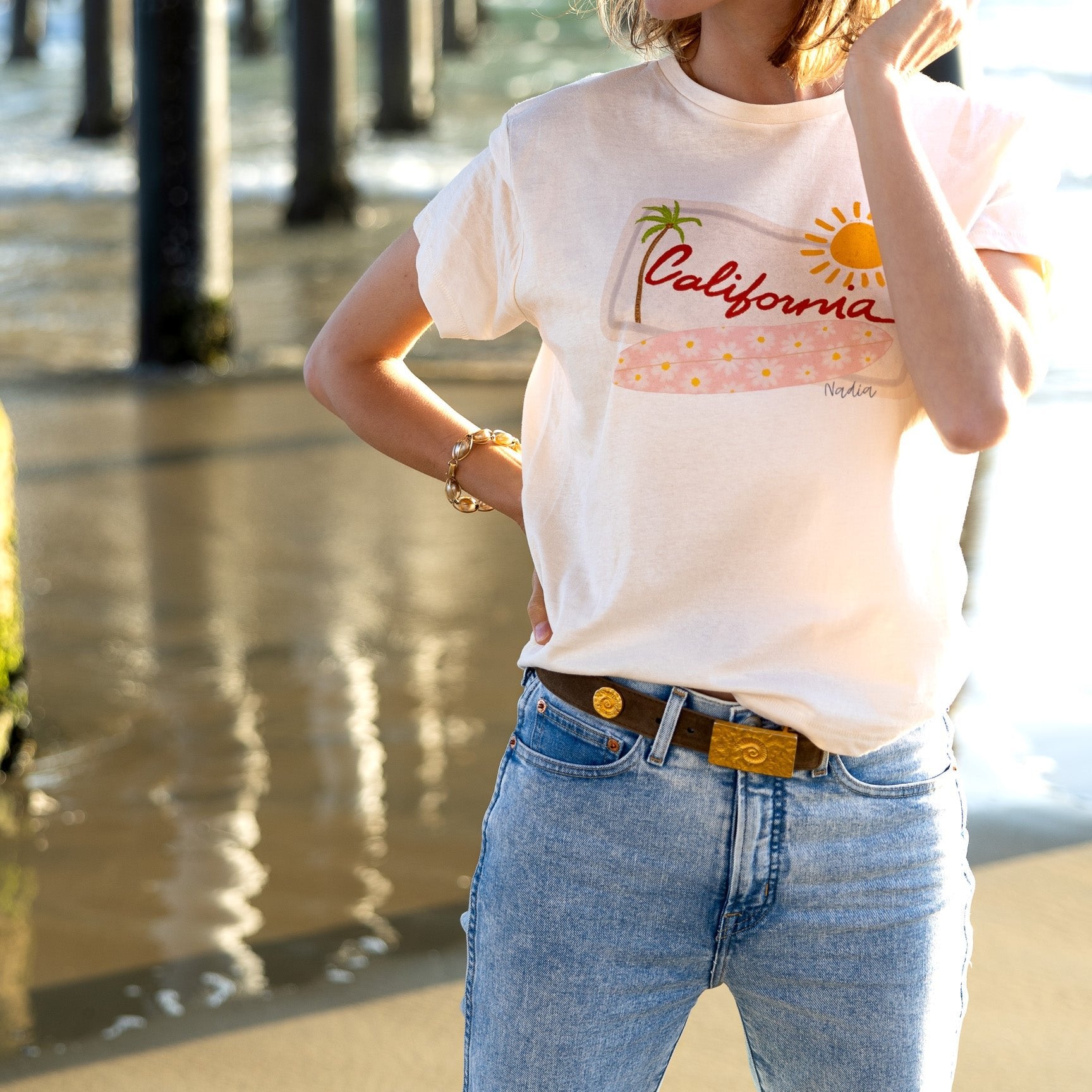 Relaxed fit women's graphic tee with hand-drawn California design, featuring a pink floral surfboard, palm tree, and sun on a white background