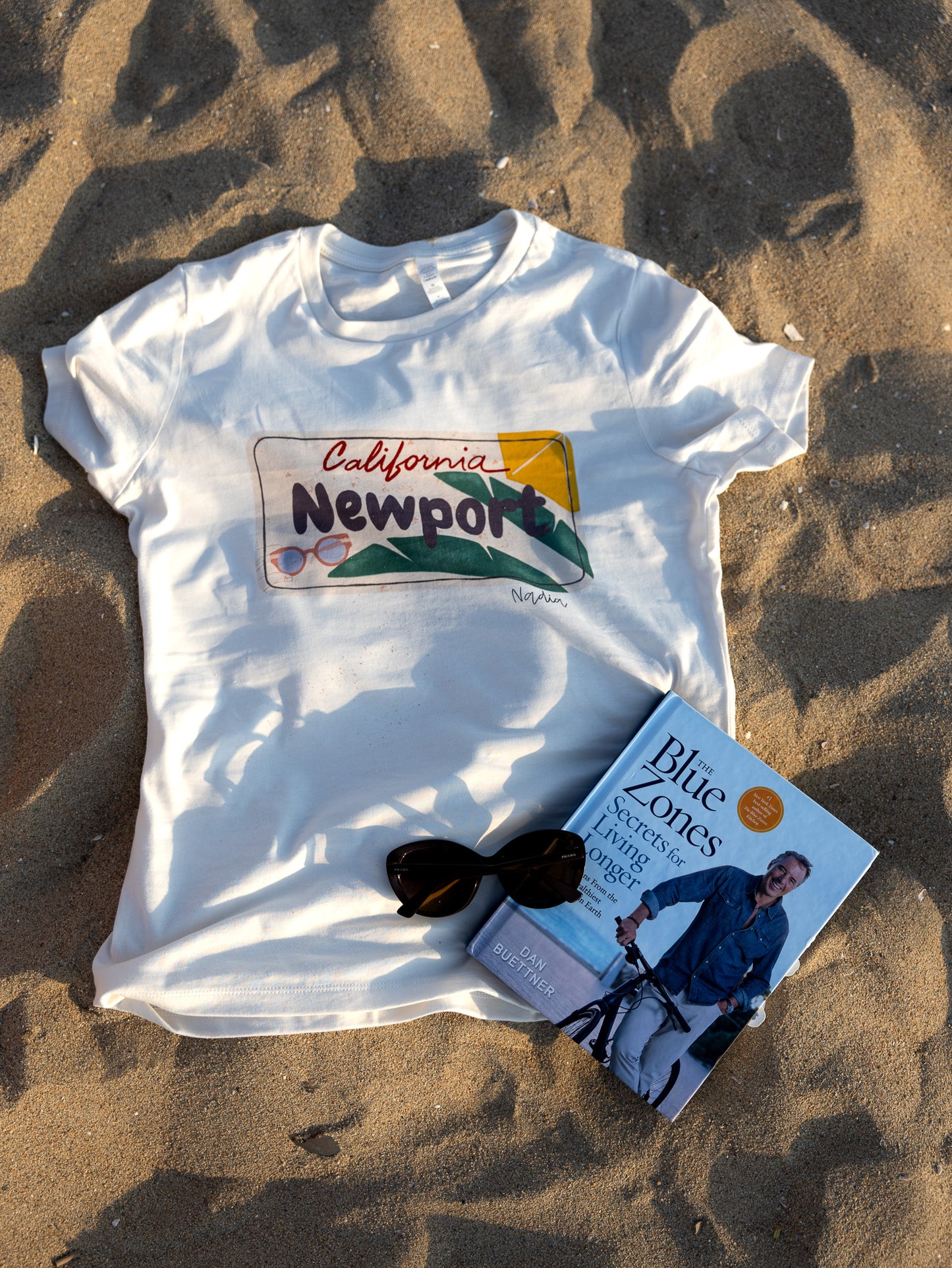 Image of a t-shirt with a hand-drawn graphic of a Newport, California license plate, showcasing a unique and comfortable way to express love for the West Coast lifestyle, specifically the Orange County (OC) region.