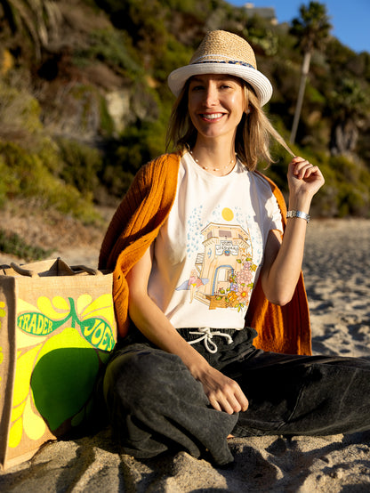 Woman wearing a cream North Shore Girls tee with an illustration of the Laguna Beach lifeguard tower and a surfer.