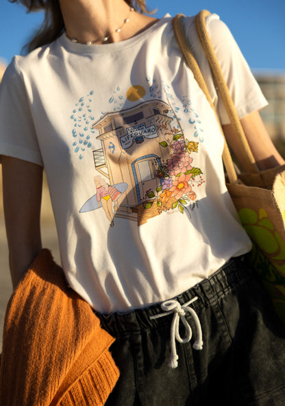 Woman wearing a cream North Shore Girls tee with an illustration of the Laguna Beach lifeguard tower and a surfer.