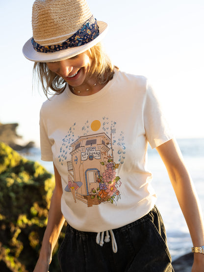 Woman wearing a cream North Shore Girls tee with an illustration of the Laguna Beach lifeguard tower and a surfer.