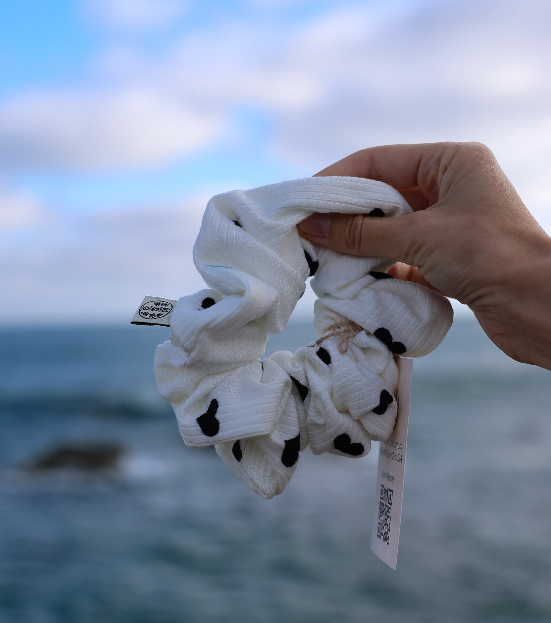 White scrunchies with black dots held by a hand against a blurred ocean and sky background