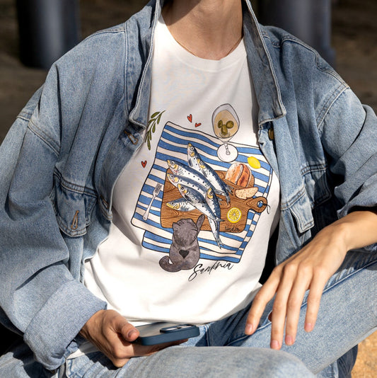 Woman is wearing a graphic tee with hand-illustrated sardines on a cutting board, dirty martini, bread, lemon, and a cute cat reaching for sardines. SARDINIA slogan