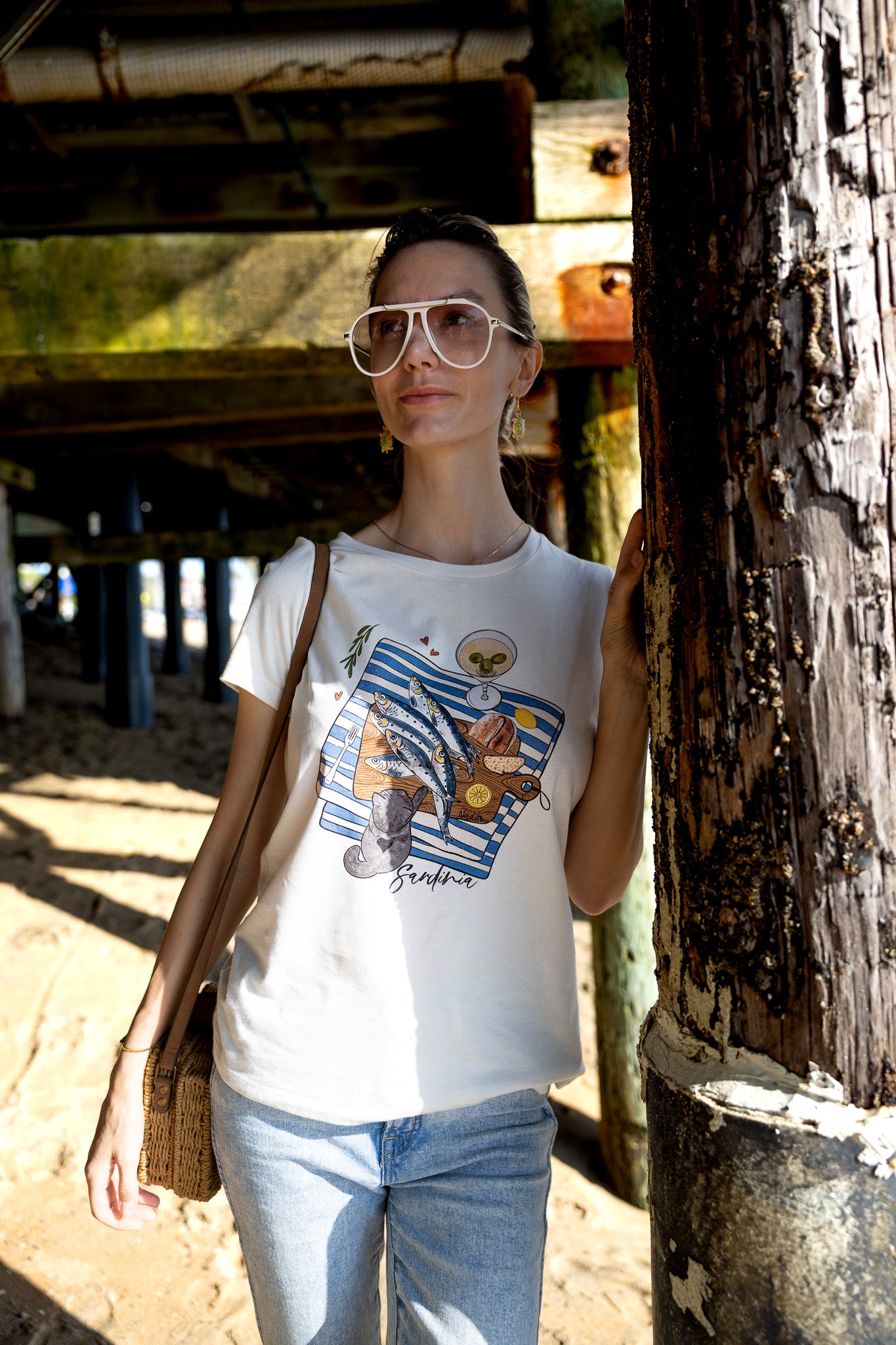 Woman is wearing a graphic tee with hand-illustrated sardines on a cutting board, dirty martini, bread, lemon, and a cute cat reaching for sardines. SARDINIA slogan