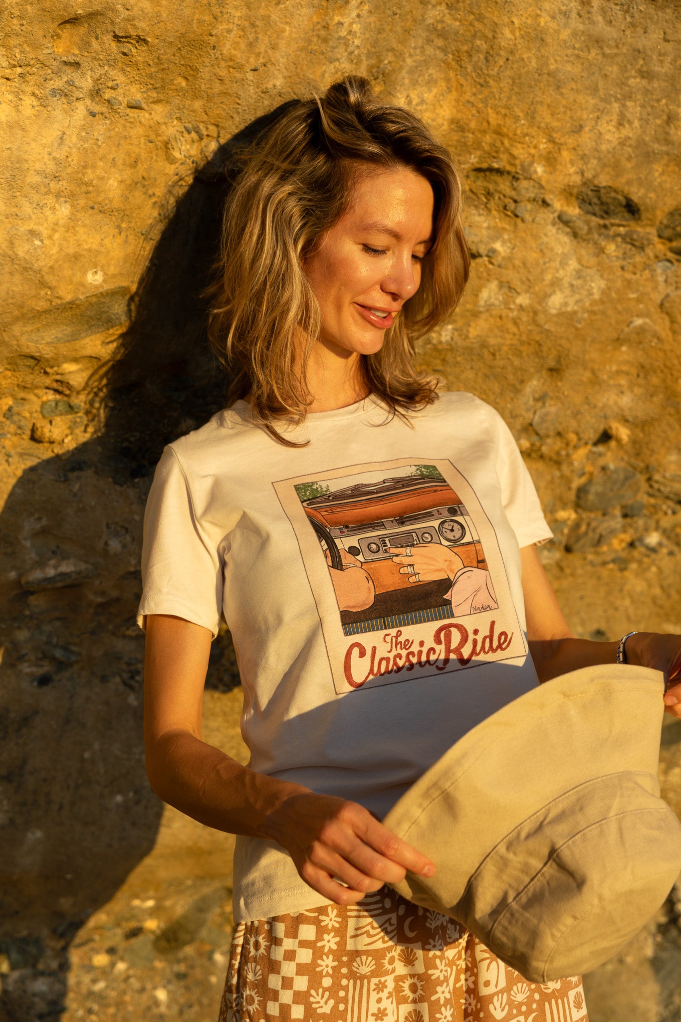 Woman wearing a t-shirt with a graphic design, holding a cap against a stone wall.