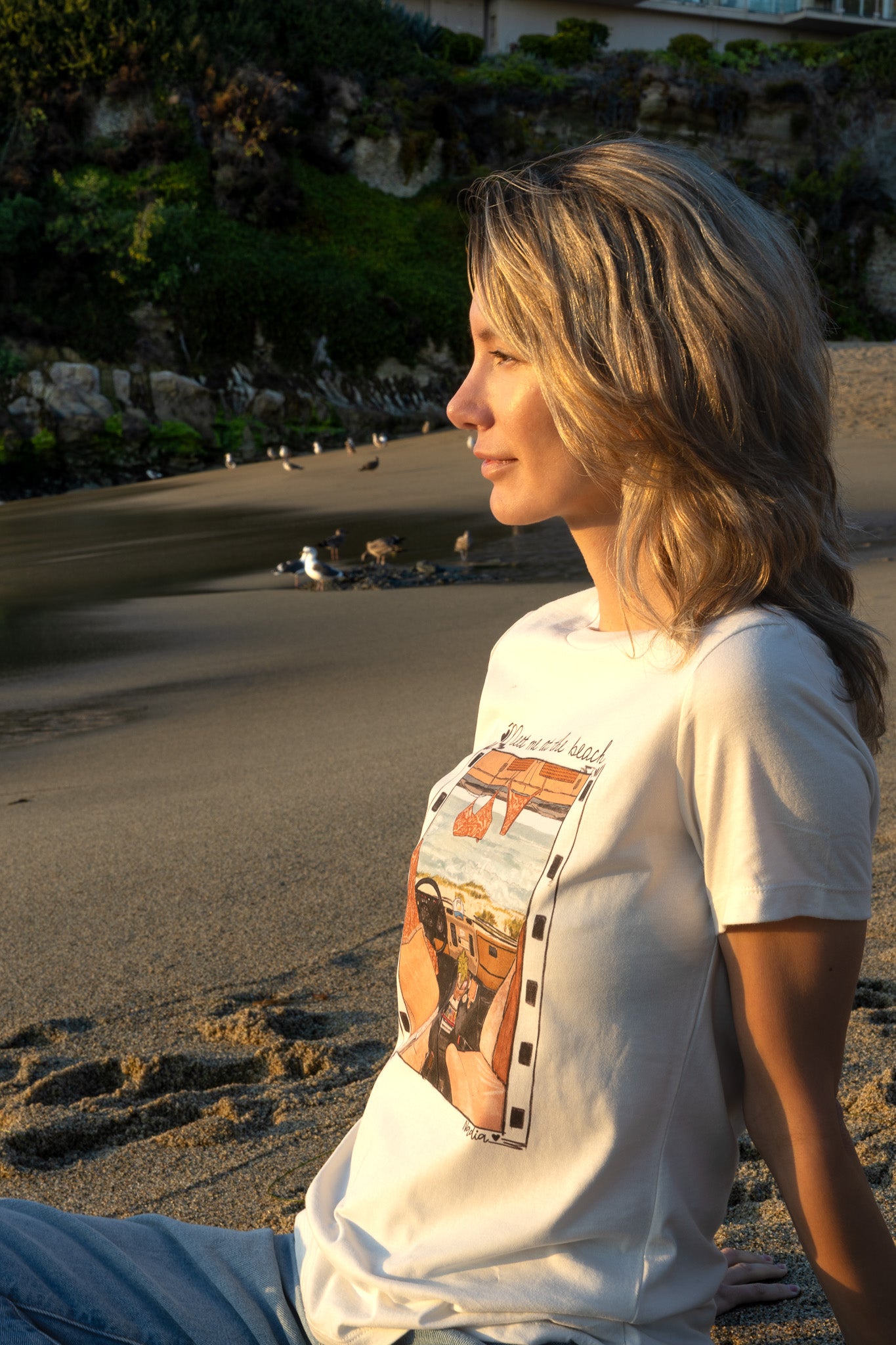 Woman sitting on a beach wearing a white t-shirt with a graphic design.