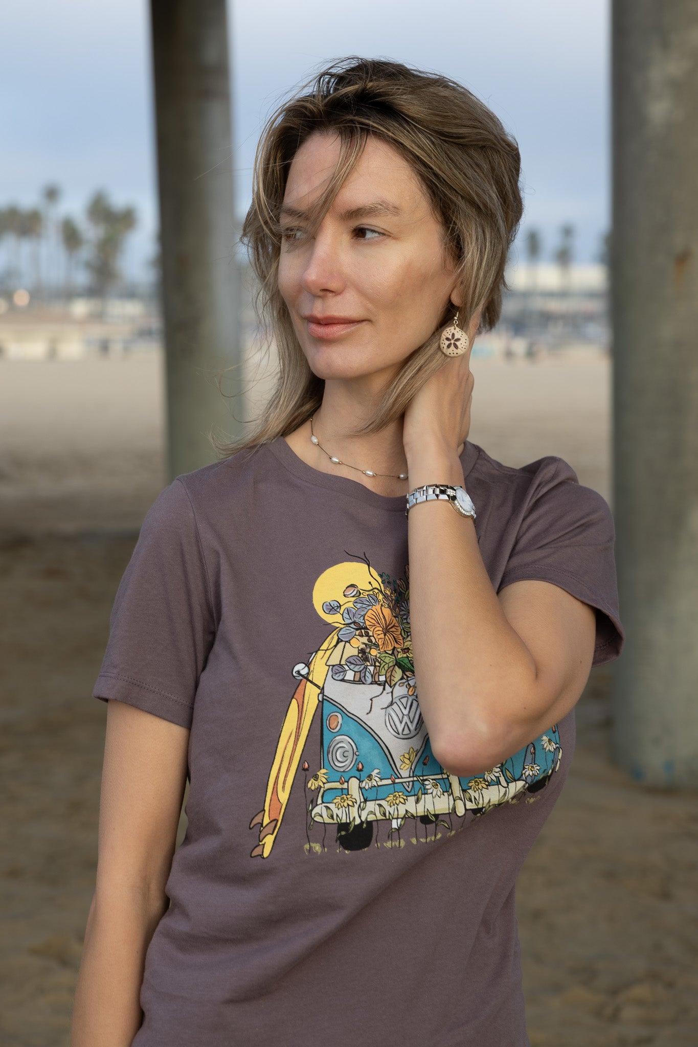 Woman wearing a graphic t-shirt with a beach scene design, standing on a sandy beach with palm trees in the background.
