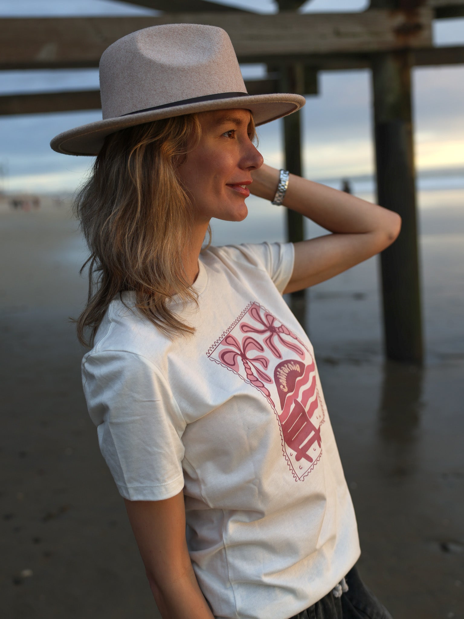 Woman wearing a white t-shirt with a red design and a beige hat on a beach.