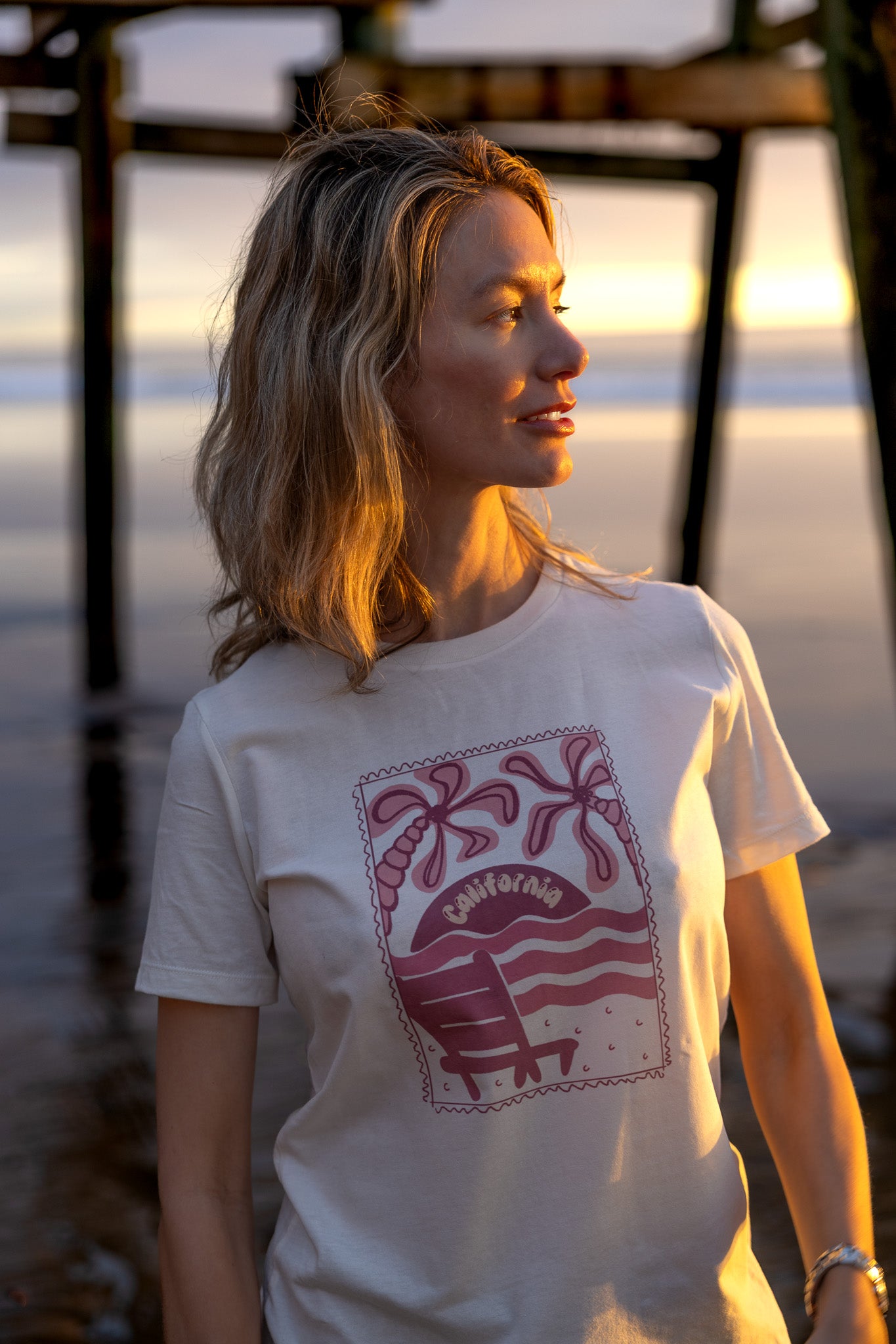 Woman wearing a white t-shirt with a red graphic design, standing by water at sunset.