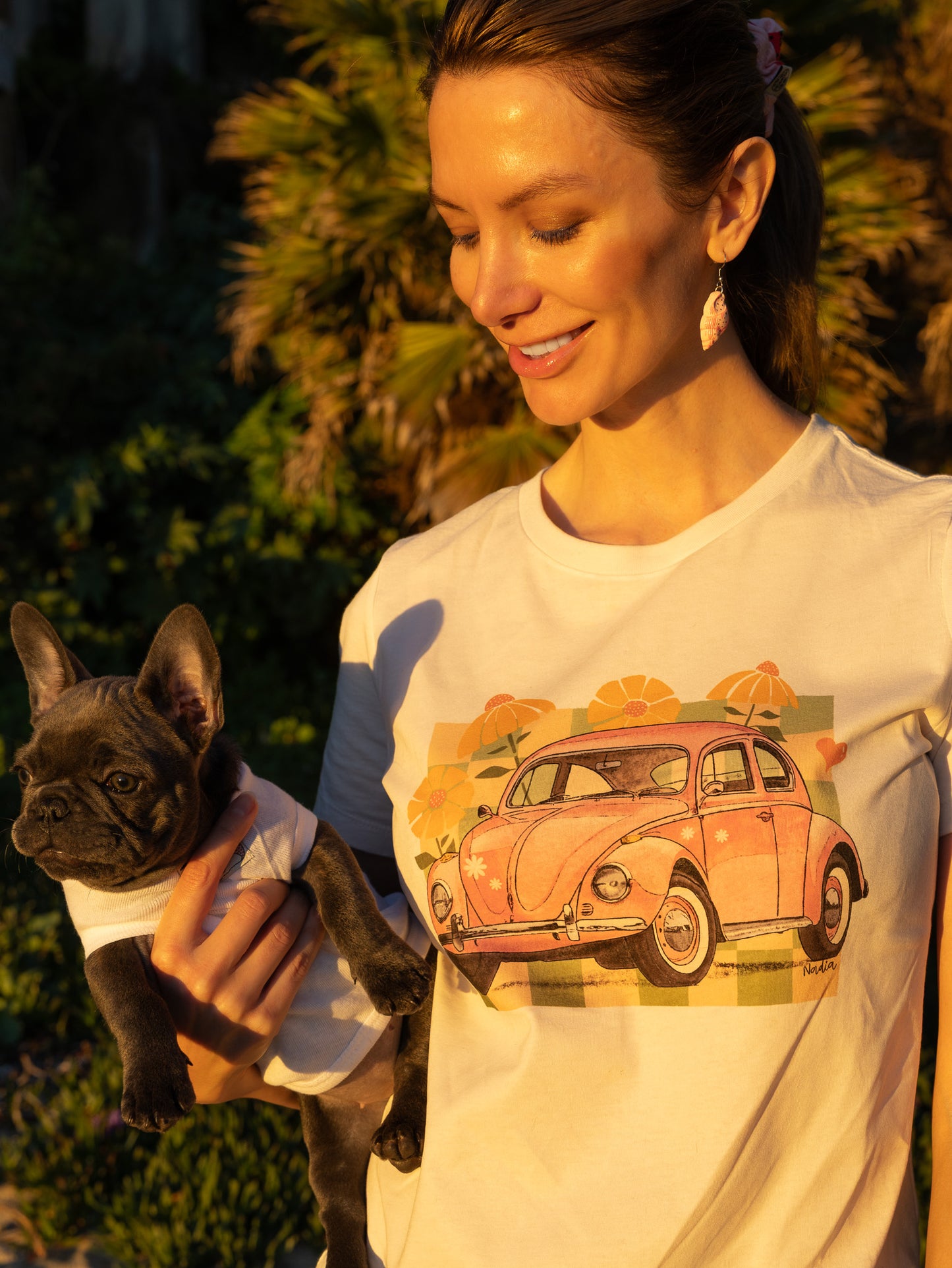 Woman standing in golden hour light wearing a cream North Shore Girls tee with a pink vintage car and checkered illustration.