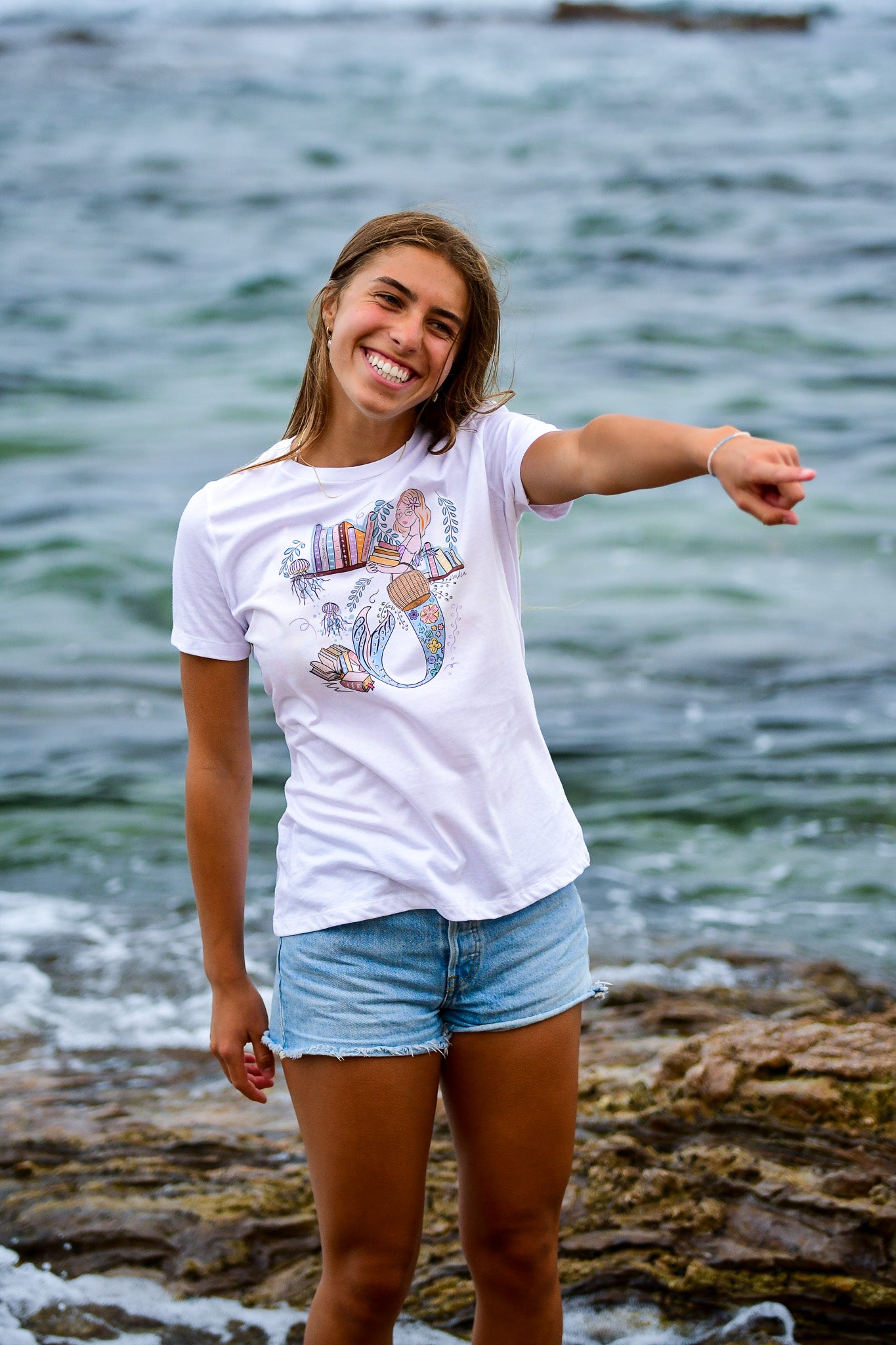 Illustrated graphic tee featuring a mermaid librarian surrounded by books and ocean elements, with a whimsical and colorful design that combines a love of reading and mythical creatures, printed on a soft and relaxed-fit cotton t-shirt.