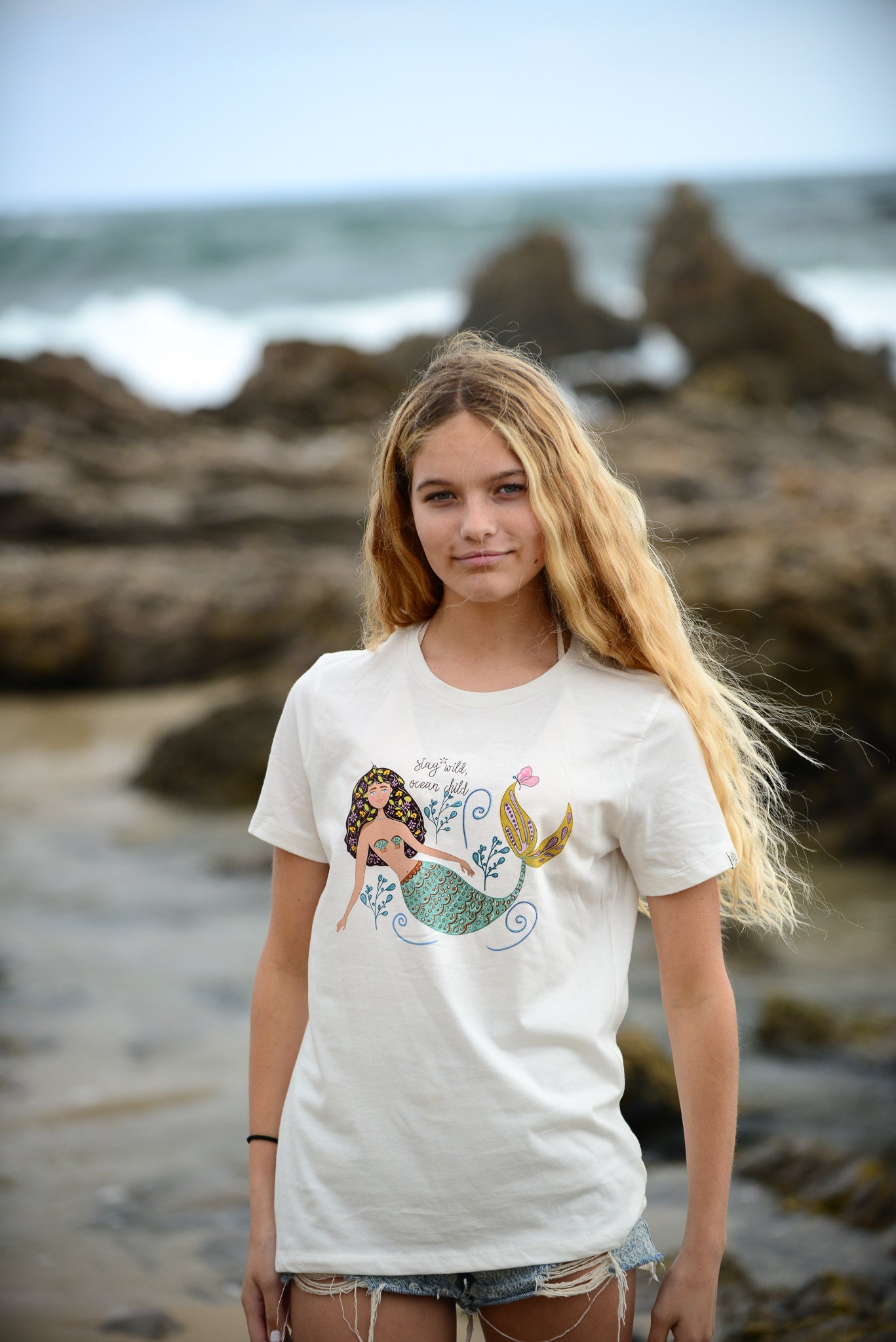 Illustrated mermaid graphic tee for women with 'Stay Wild Ocean Child' slogan. A whimsical mermaid tail and sea flowers surround the text, creating a dreamy ocean-inspired design on a relaxed-fit cotton t-shirt.