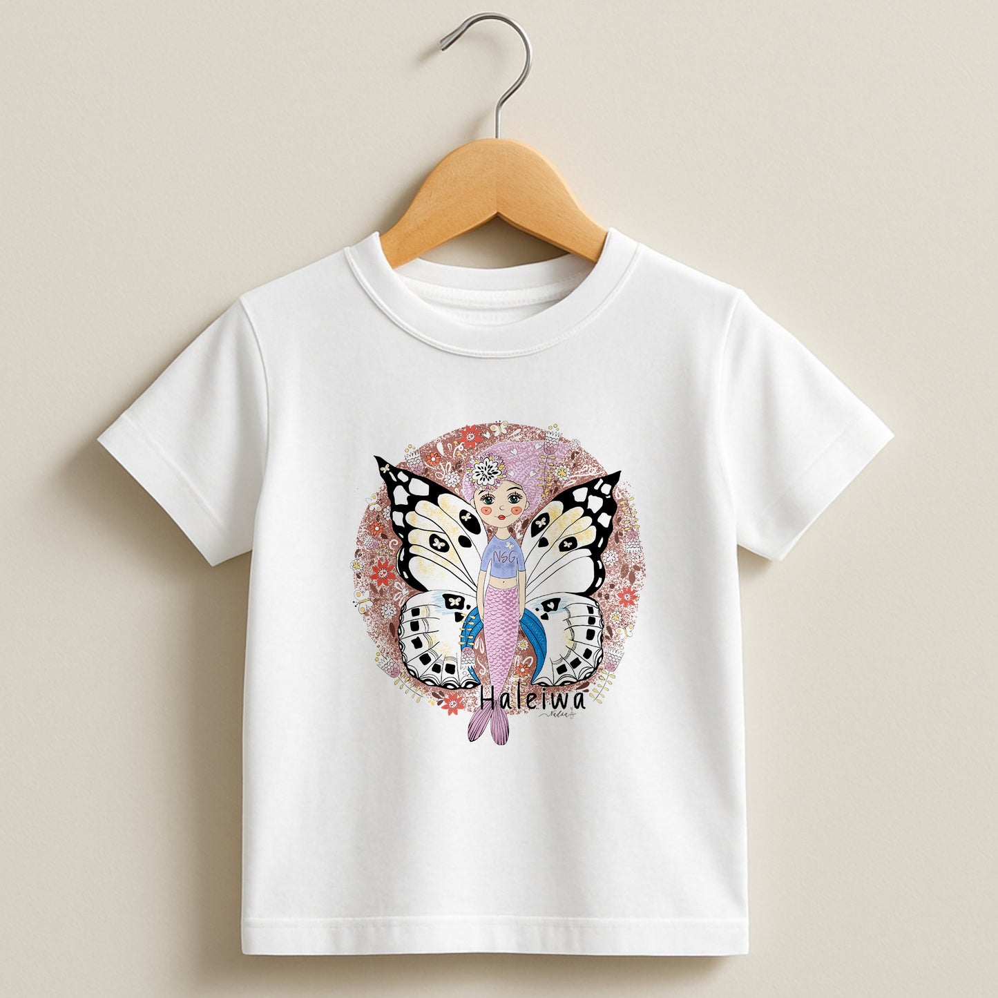 White toddler t-shirt featuring a hand-drawn mermaid with butterfly wings and floral patterns, labeled Haleiwa.