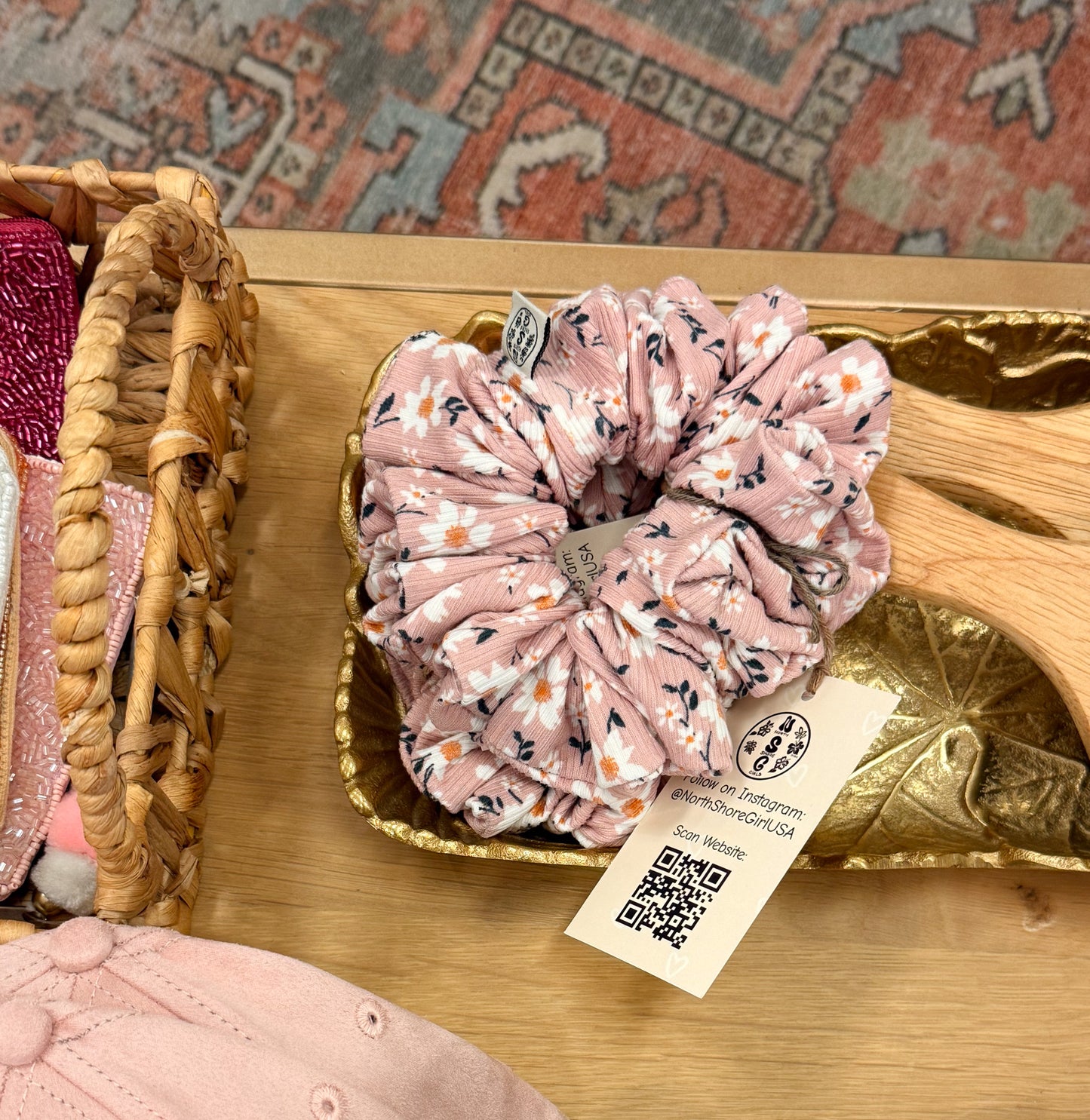 Pink floral scrunchie in a gold container with a tag on a wooden surface.
