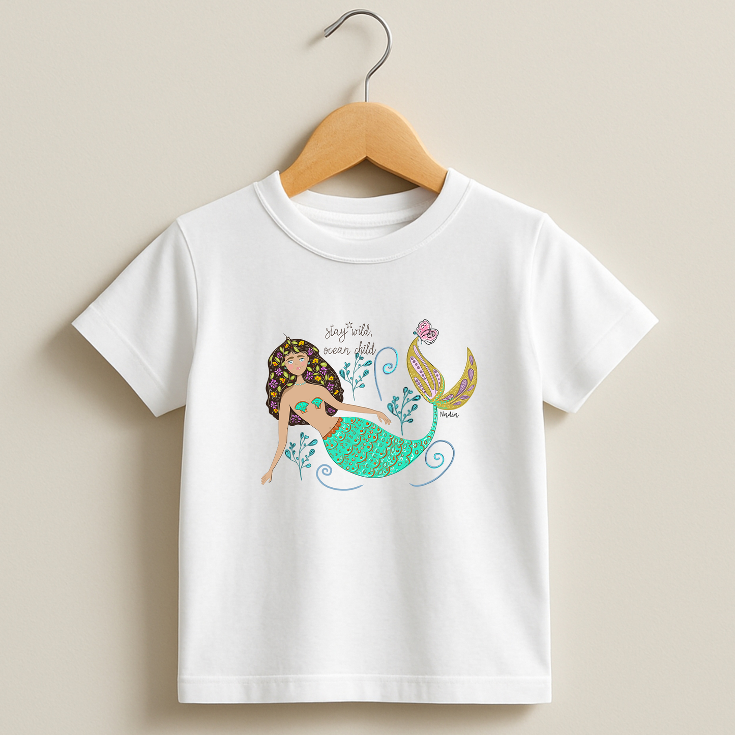 a white tee with a colorful mermaid illustration and “Stay Wild Ocean Child” slogan – original artwork by Nadia Watts