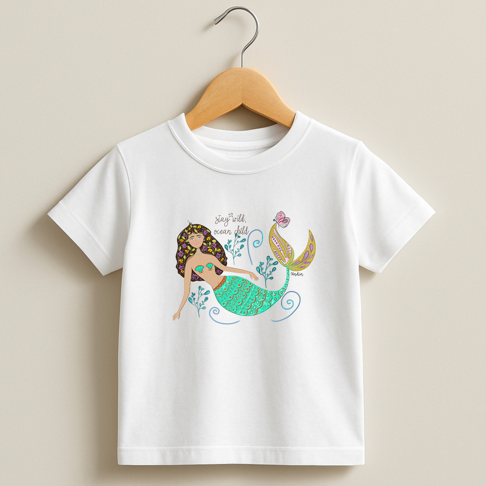 a white tee with a colorful mermaid illustration and “Stay Wild Ocean Child” slogan – original artwork by Nadia Watts