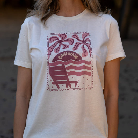 Women's cream-colored tee with a mauve postage stamp illustration of a beach chair and palms.