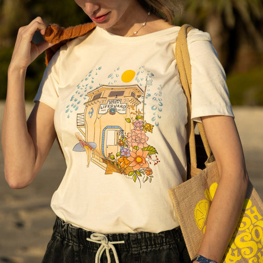 Woman wearing a cream North Shore Girls tee with an illustration of the Laguna Beach lifeguard tower and a surfer.