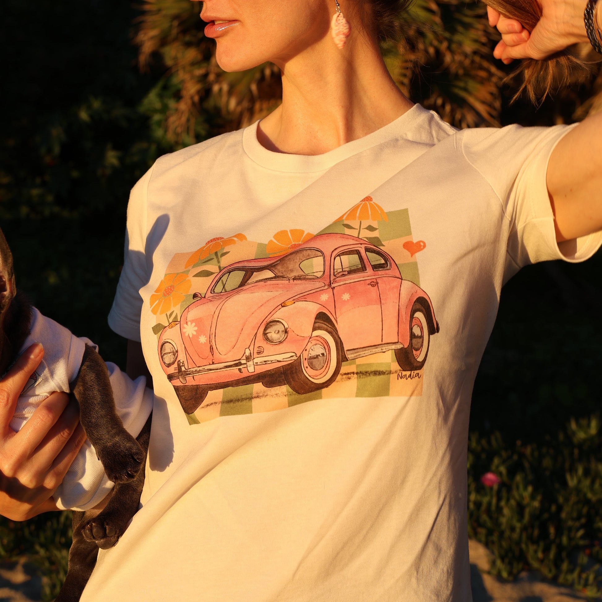Woman standing in golden hour light wearing a cream North Shore Girls tee with a pink vintage car and checkered illustration.