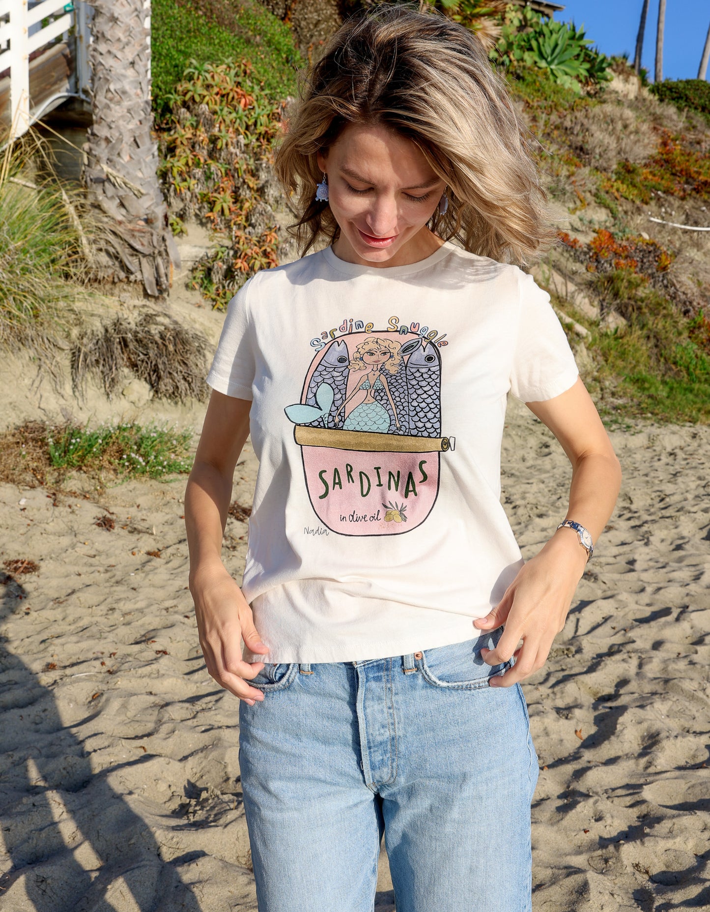 Woman standing on a beach wearing a North Shore Girls cream-colored tee with a mermaid sardine tin illustration